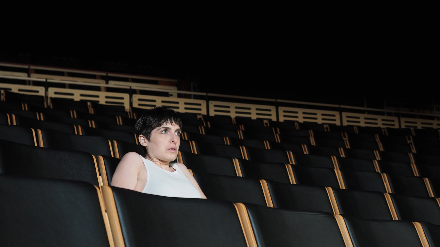 A woman wearing a white tank top sitting in a theater alone, staring at the stage with wide eyes. 