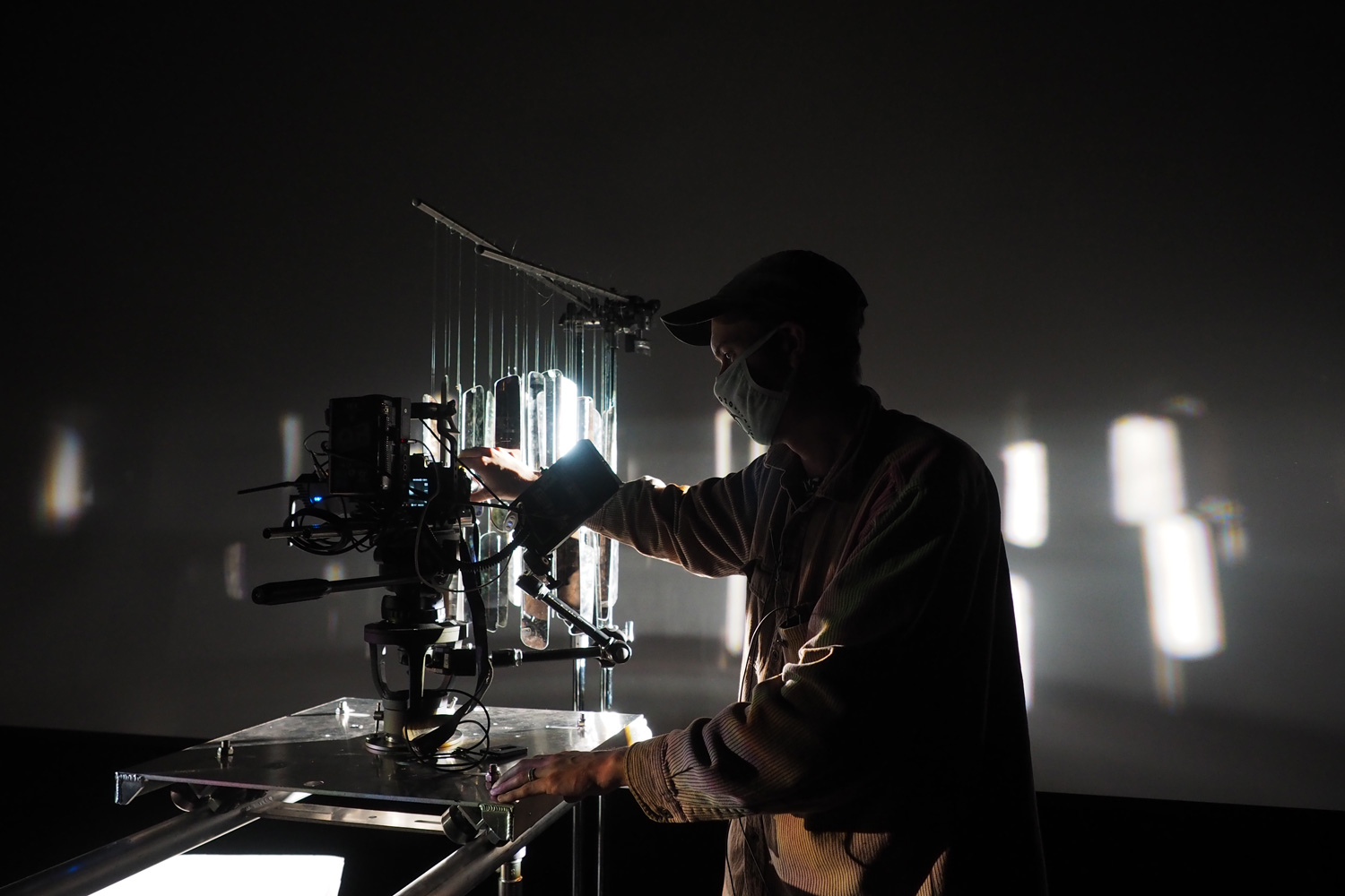 A masked production technician working with a camera stand filming a backlit wind chime. 