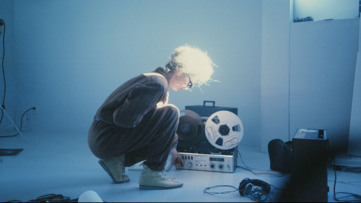 Maryanne Amacher kneeling in front of a 70's film projector in a cooly lit room.  