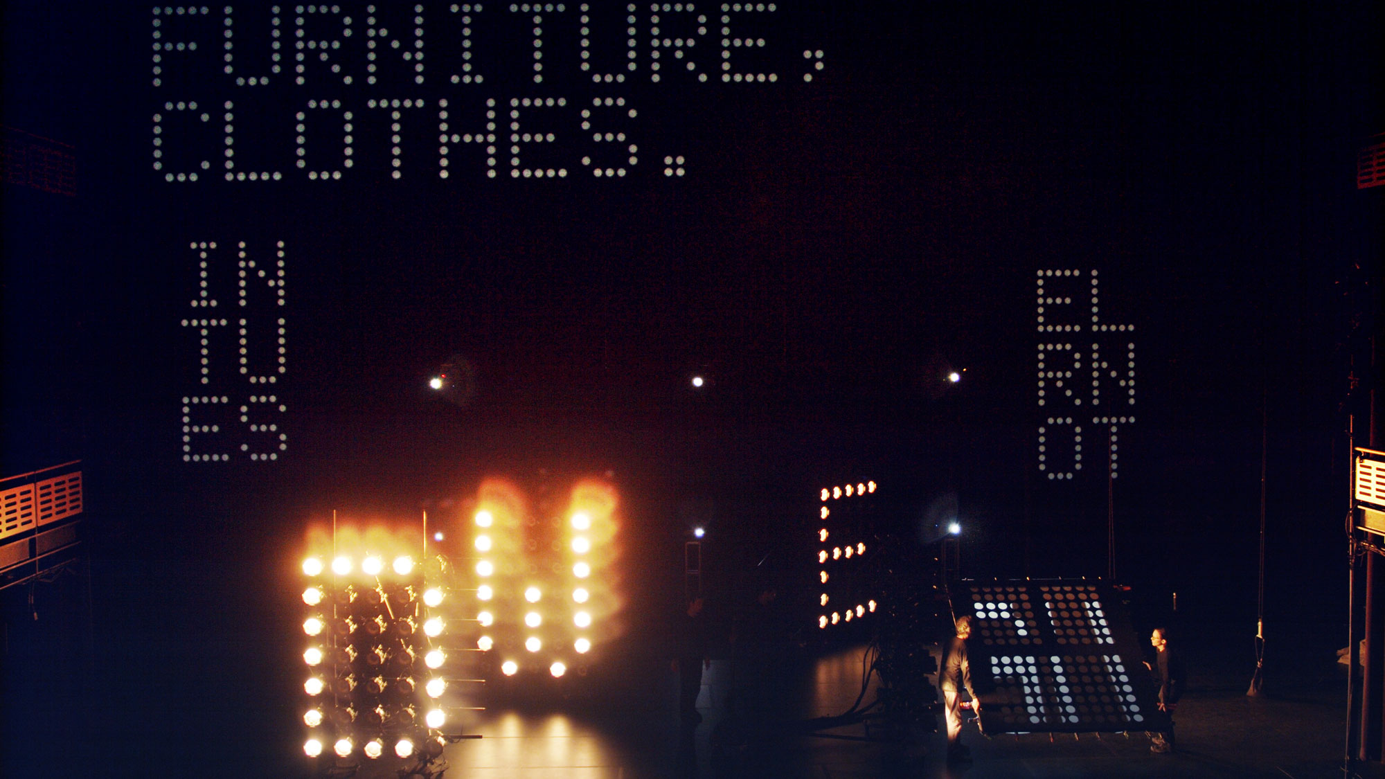words spelled out by lights in a large black studio: furniture, clothes. in tu es.