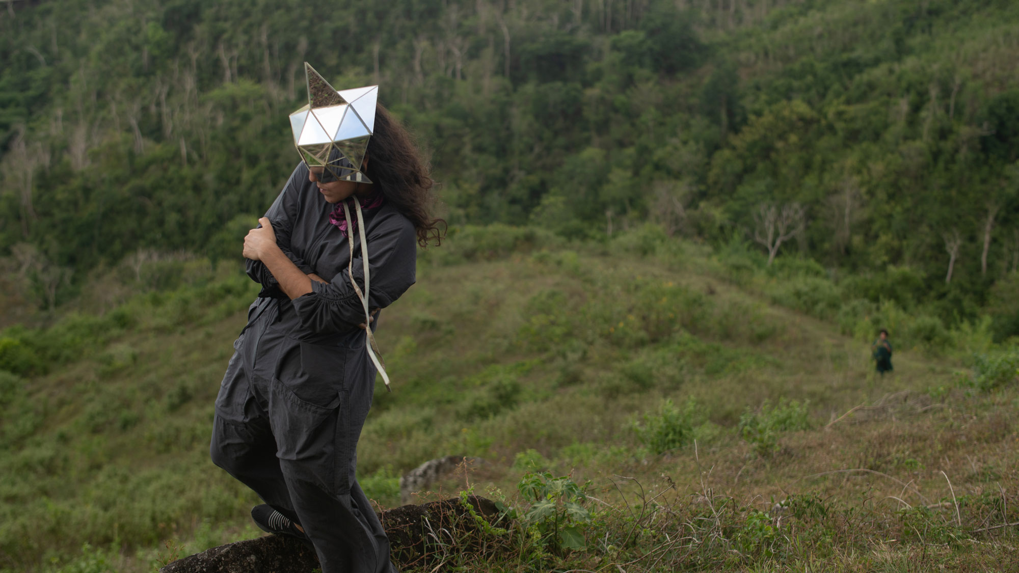 a woman in a jumpsuit with a mirrored mask standing on a precipice above a jungle
