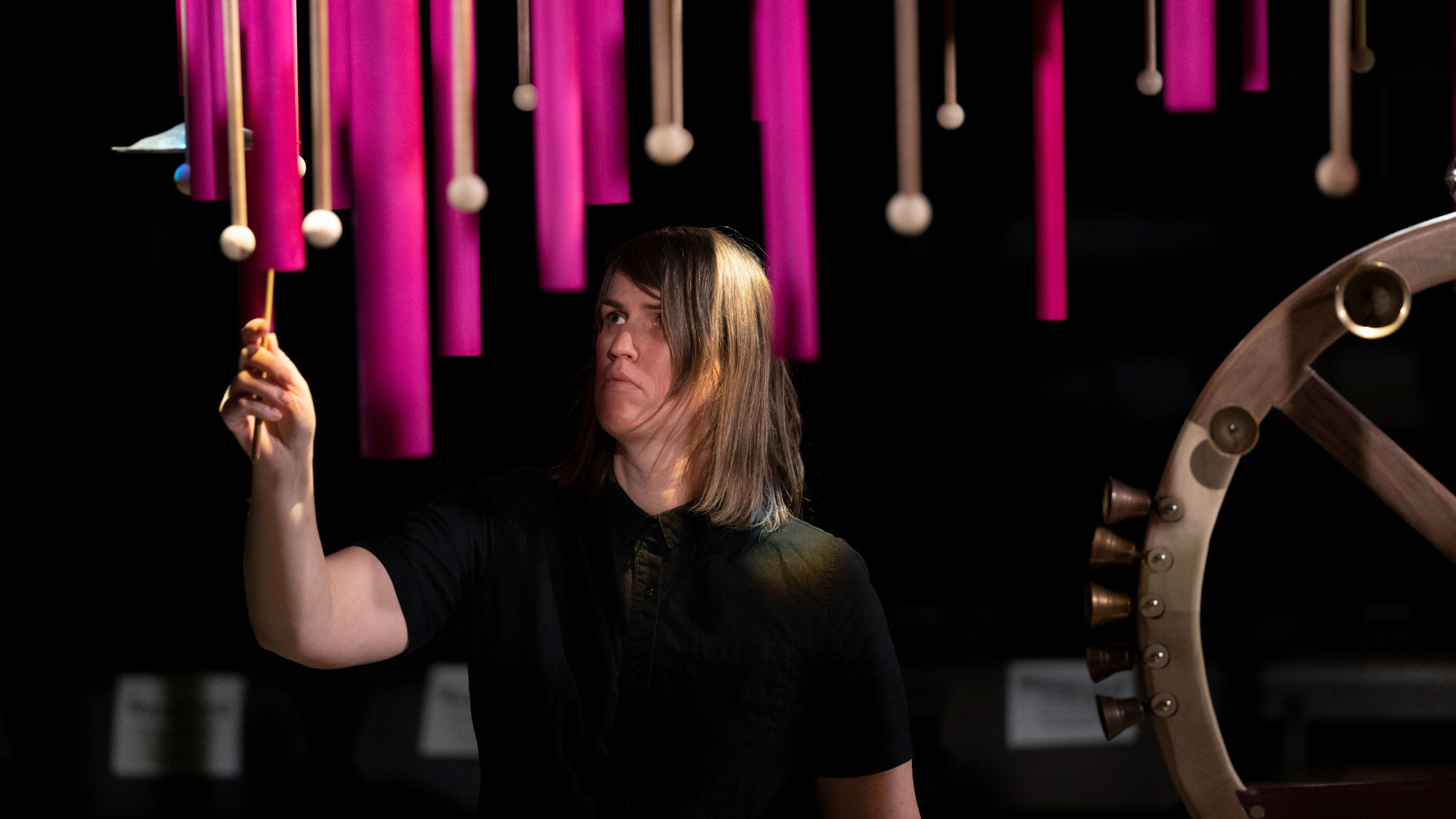 a white person in a black tshirt stands on stage with a mallet gesturing to play one hanging down from above