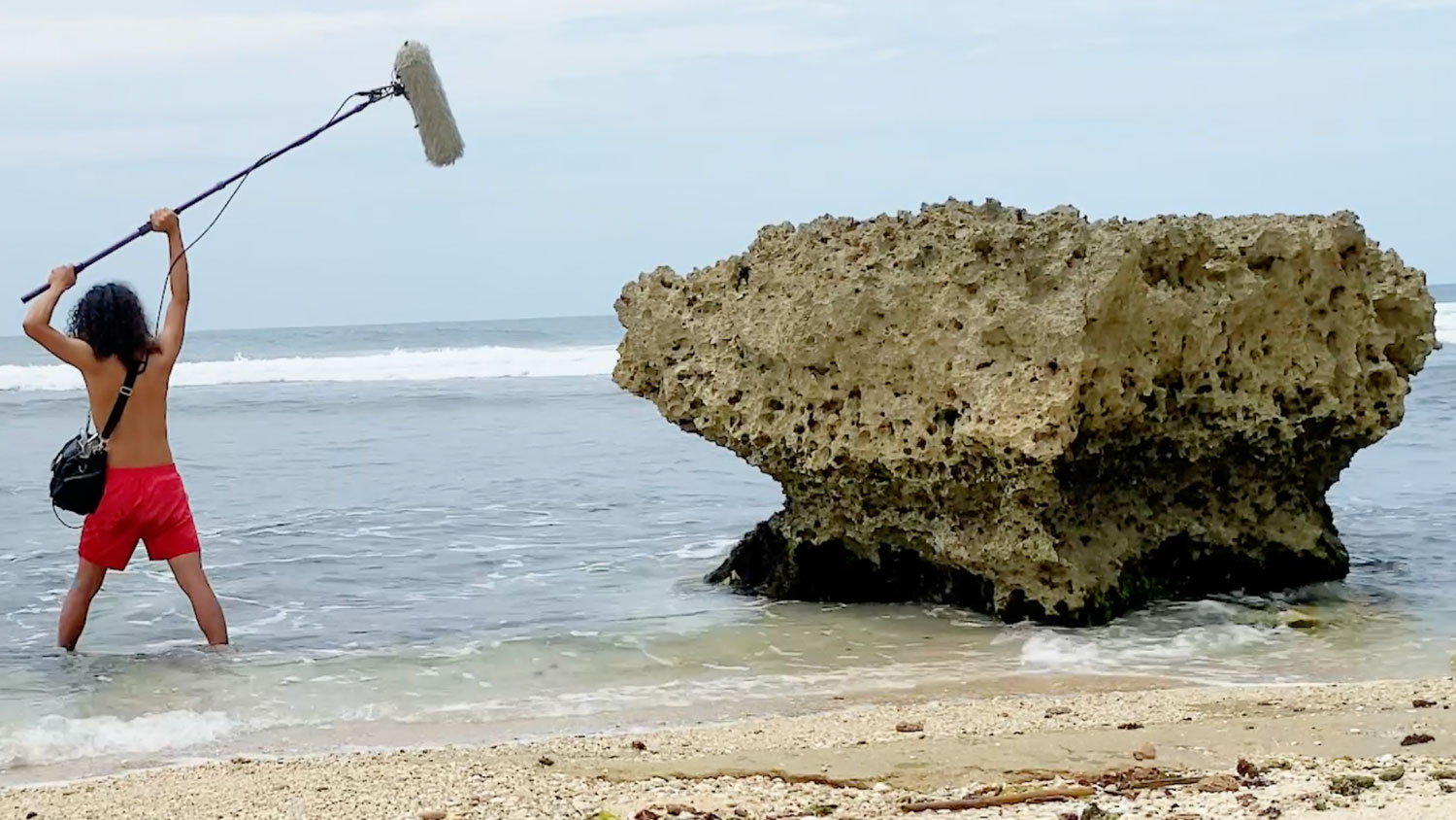 a shirtless person holding a boom microphone above the surf near a rock covered in barnacles