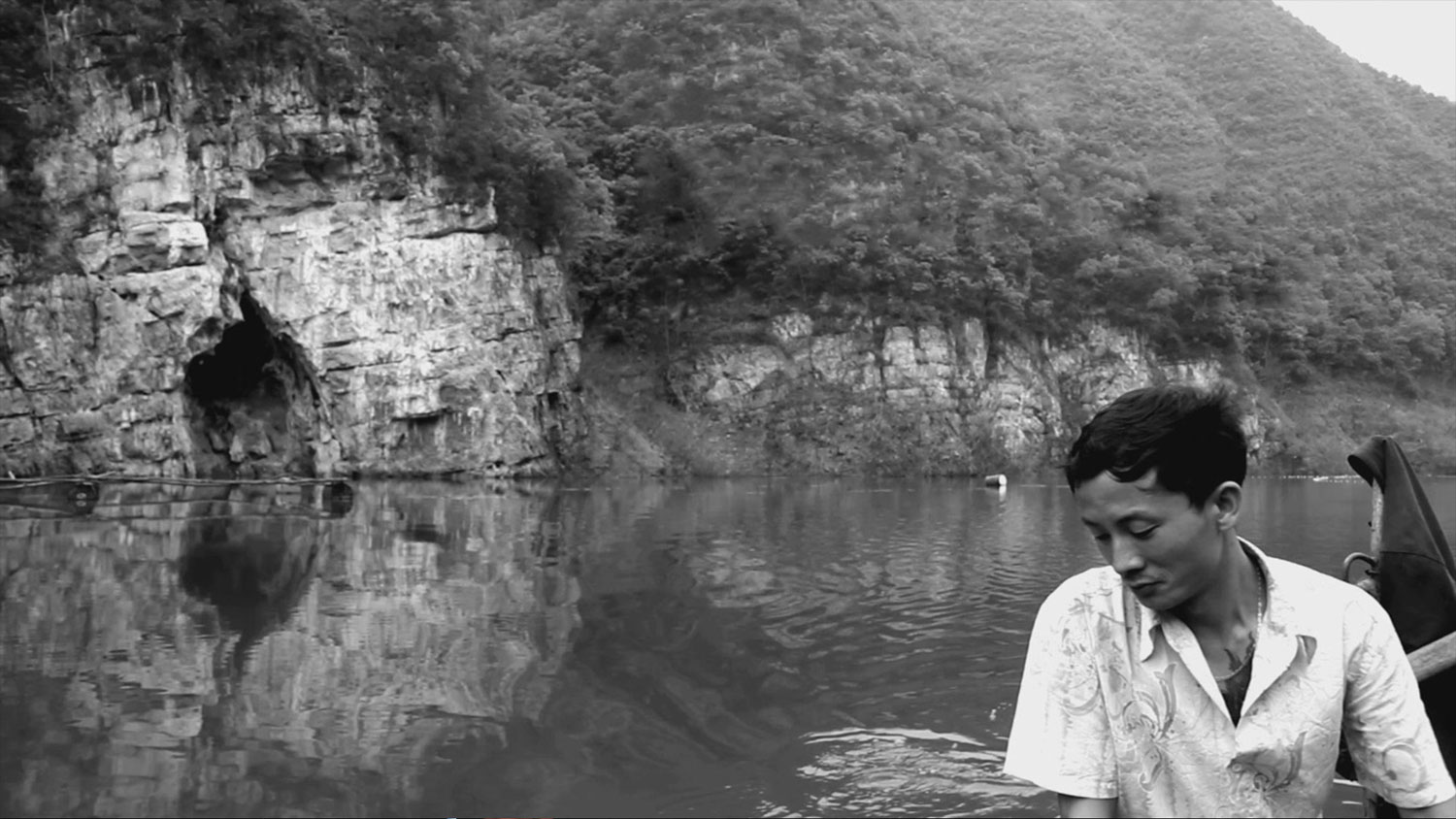 an asian person sits eyes cast down in a boat on a still lake with sheer rock sides