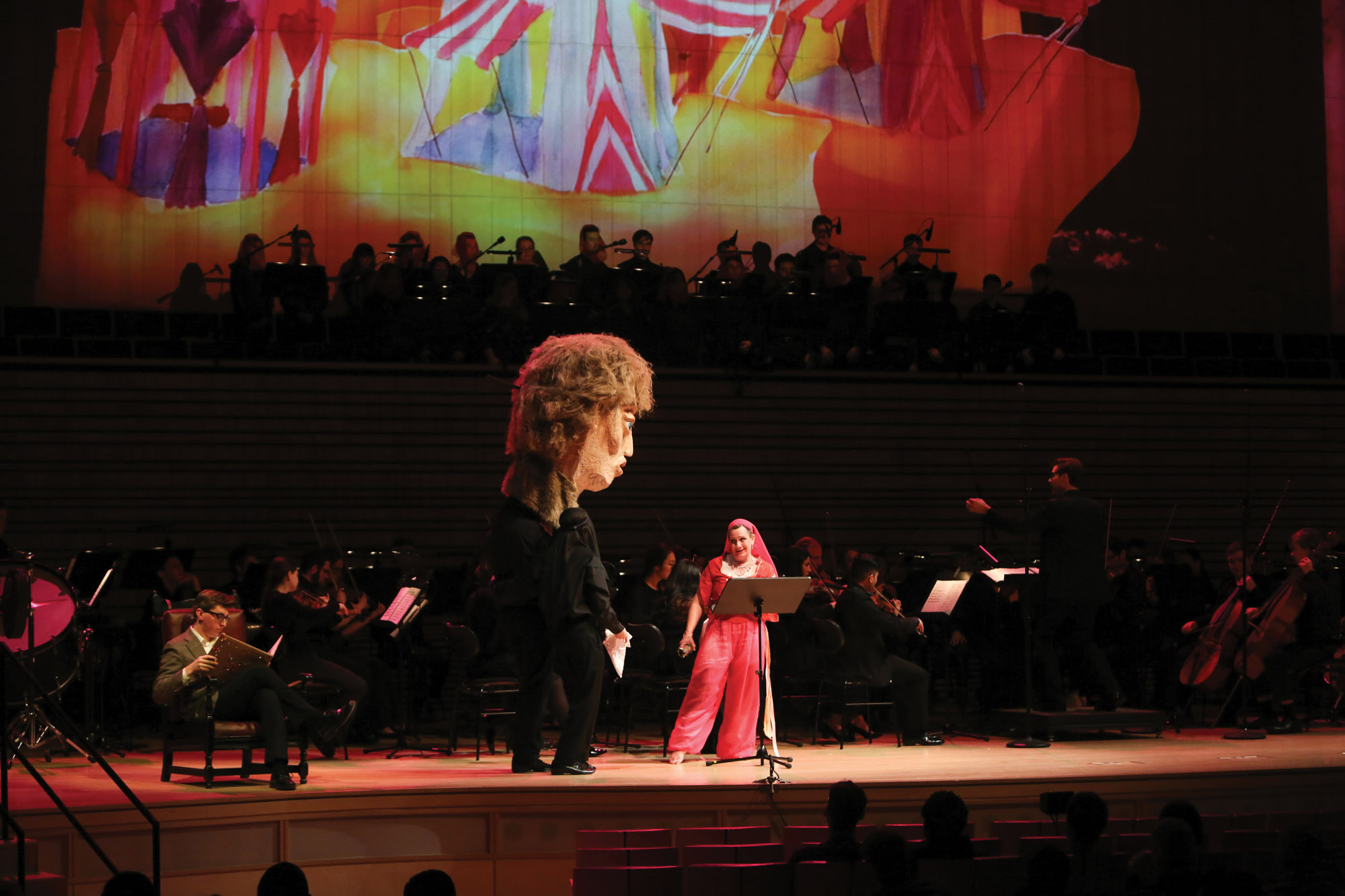 a woman dressed an a red genie costume speaking to a huge paper maché head on stage in front of a full orchestra in the EMPAC concert hall. 