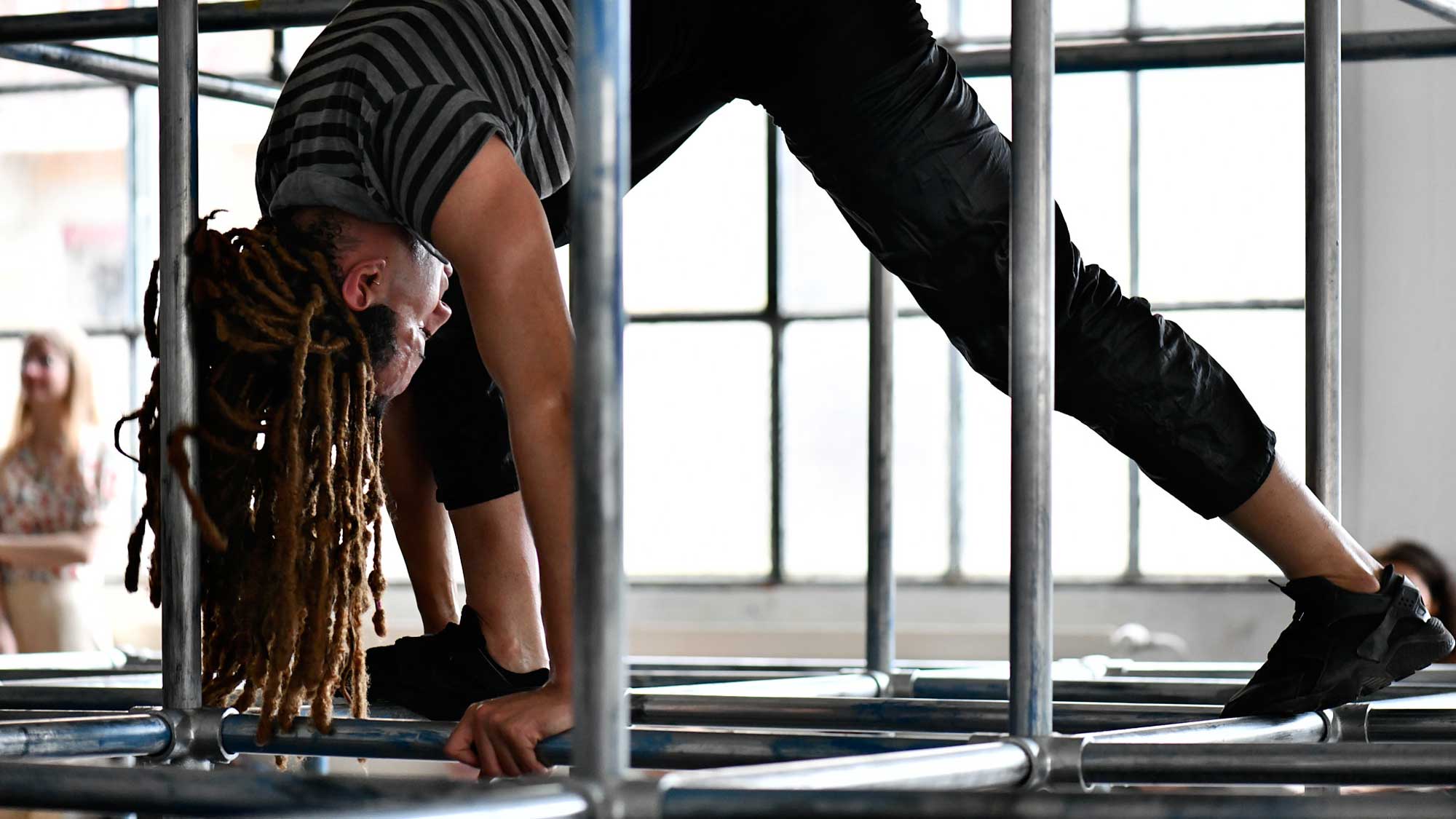 a Black performer in a galvanized steel playground-like structure bent over on the bars