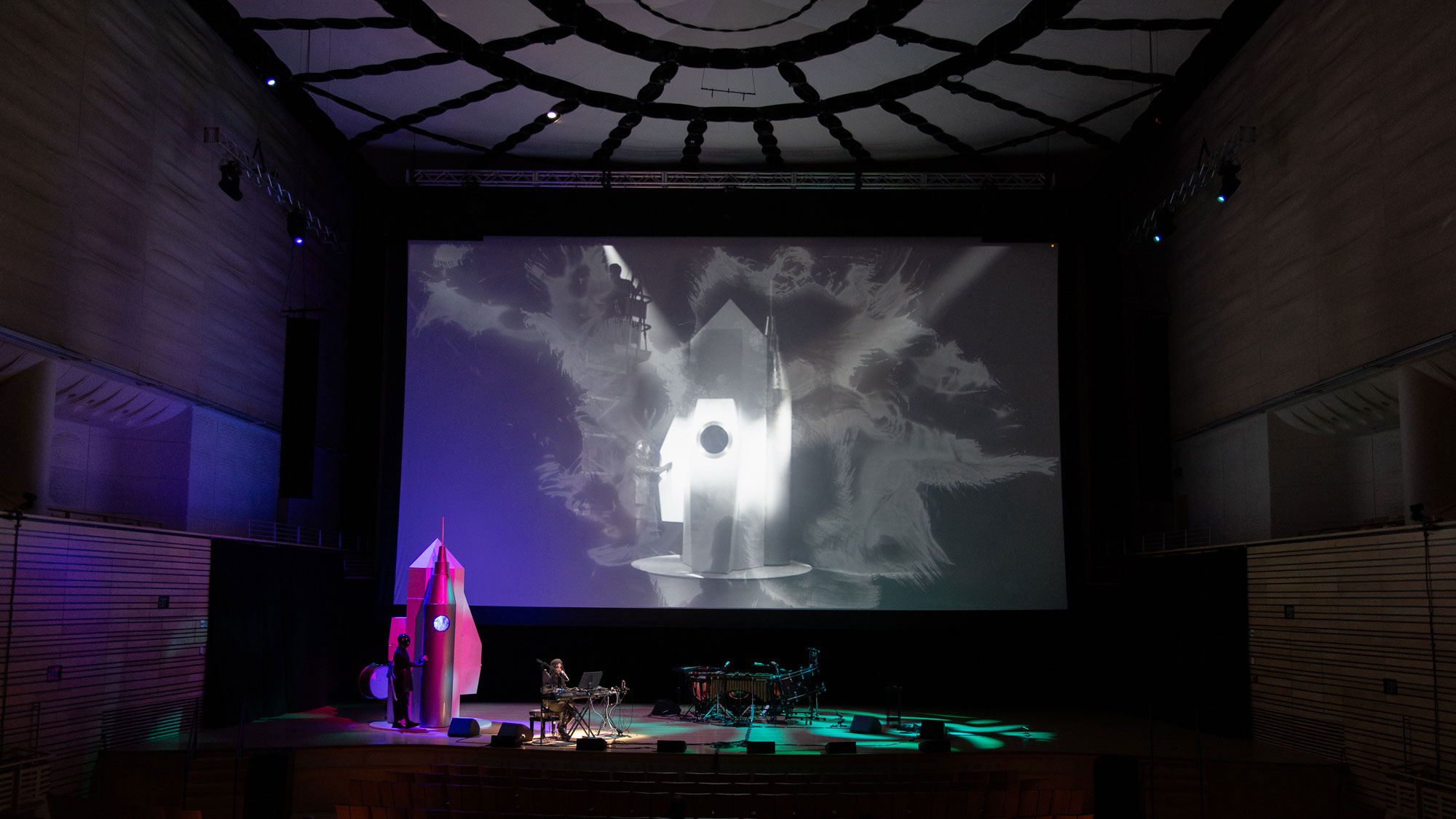 a film projected in the concert hall, a rocket ship cast in purple, mlamar performing on stage with keyboards.