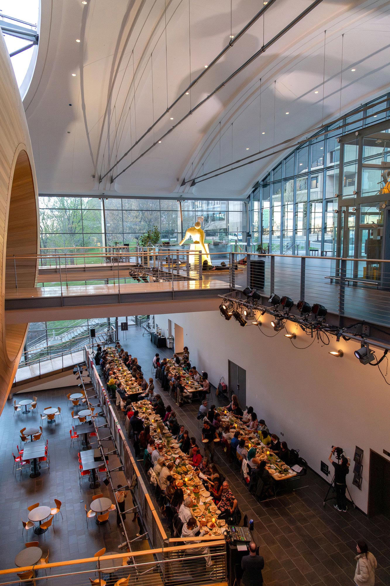 a long table meal on the mezzanine with an blow up amorphus human on a monument pedestal on the floor above it in a performing arts center