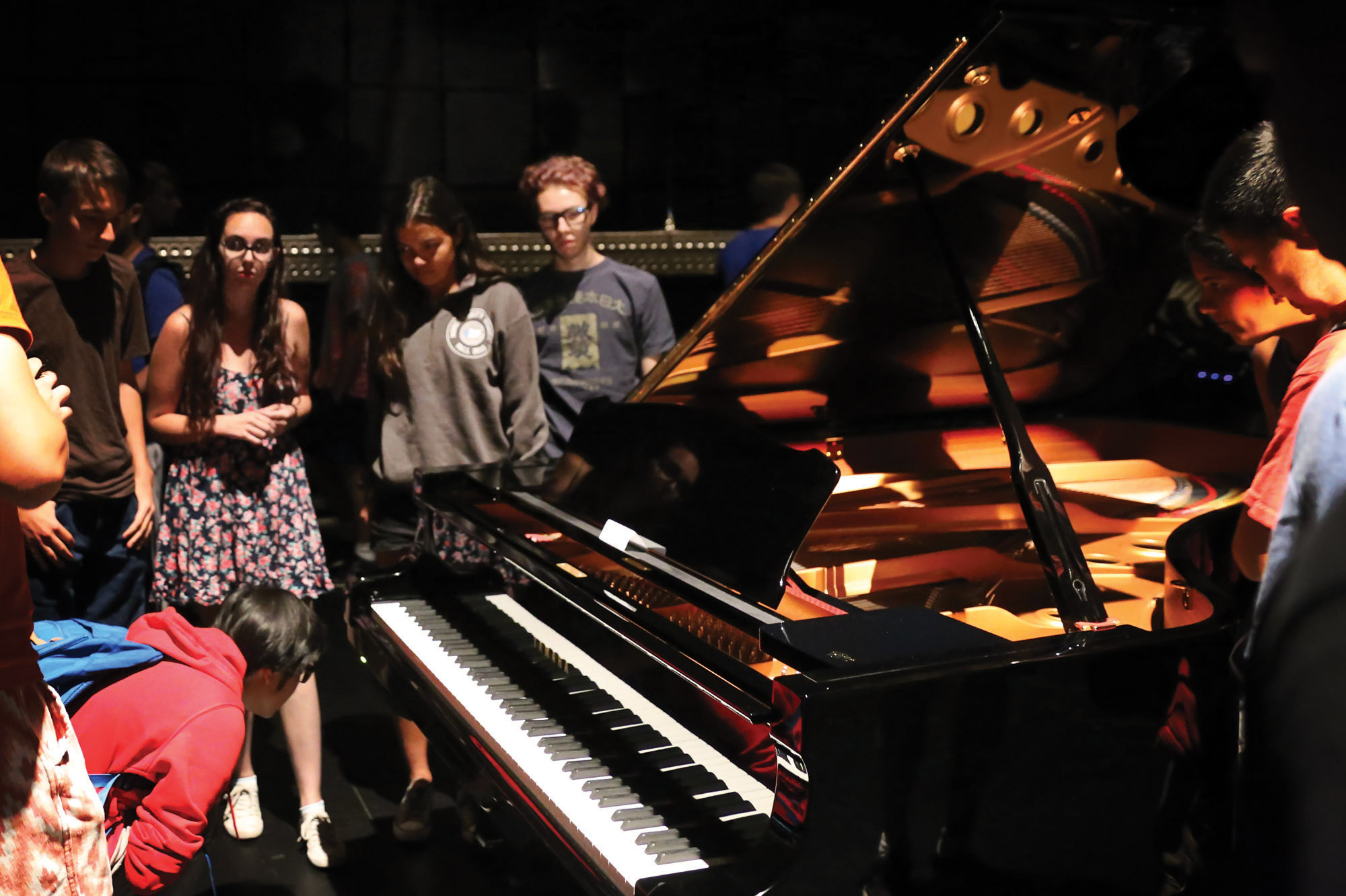 A group of students standing around a grand piano examining the keys. 