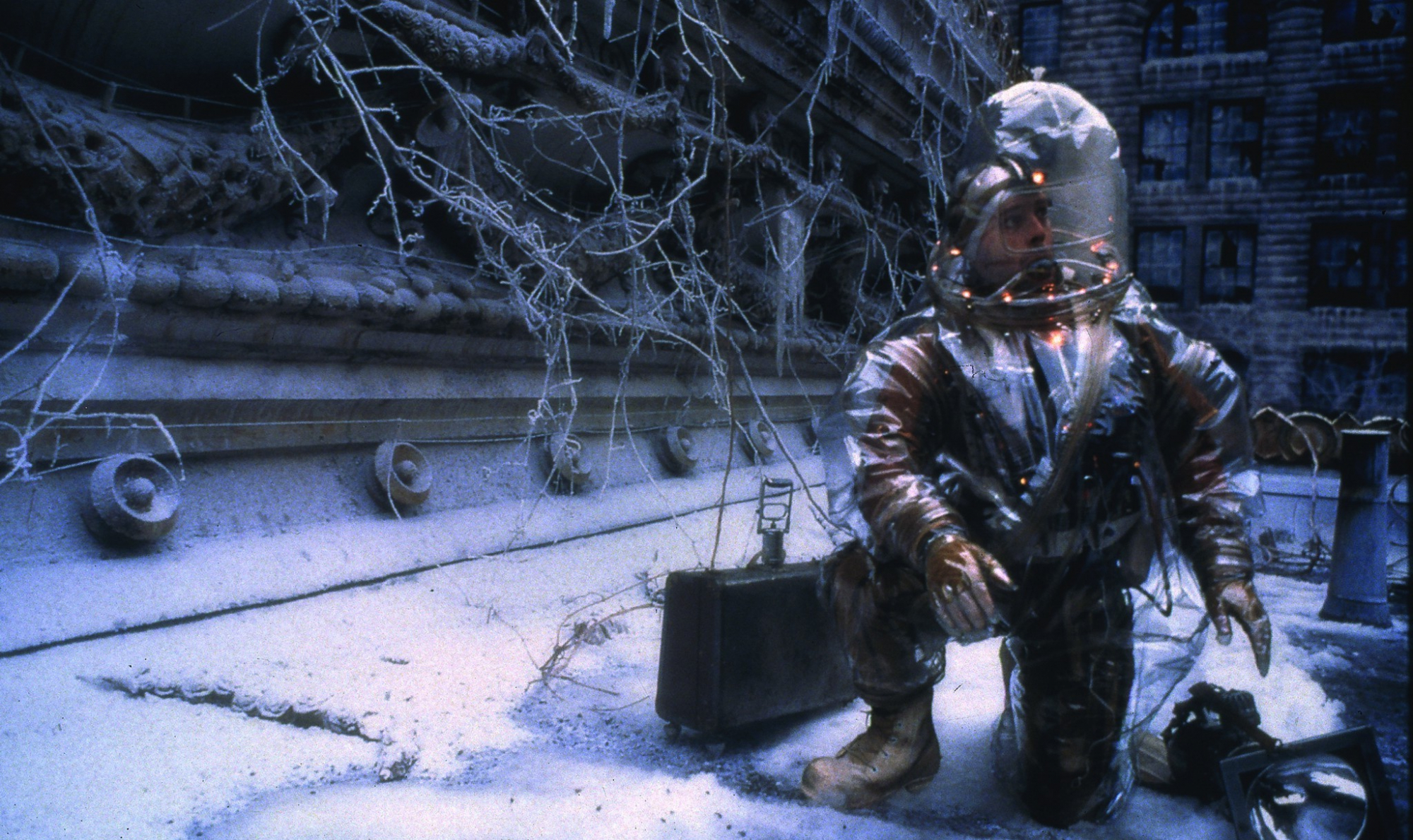 Bruce Willis kneels in a environmental hazmat suit in the snow in the film, 12 monkeys. 
