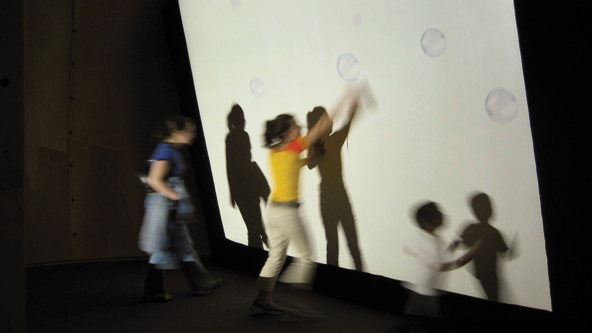 Three children blurred in motion trying to catch projected bubbles on a white wall. 