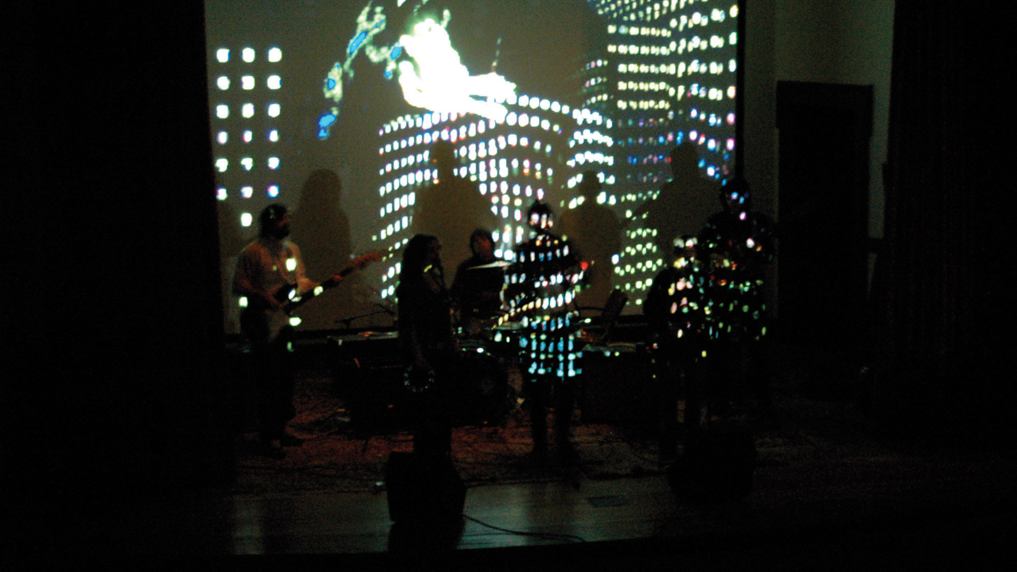 A projection of city lights against musicians and people mingling in a black box studio. 