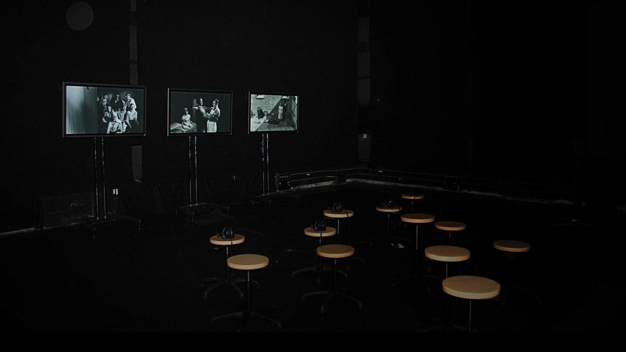 A dark black box theater with three small screens showing black and white images. Twelve round tan stools are are set up in front of them. 