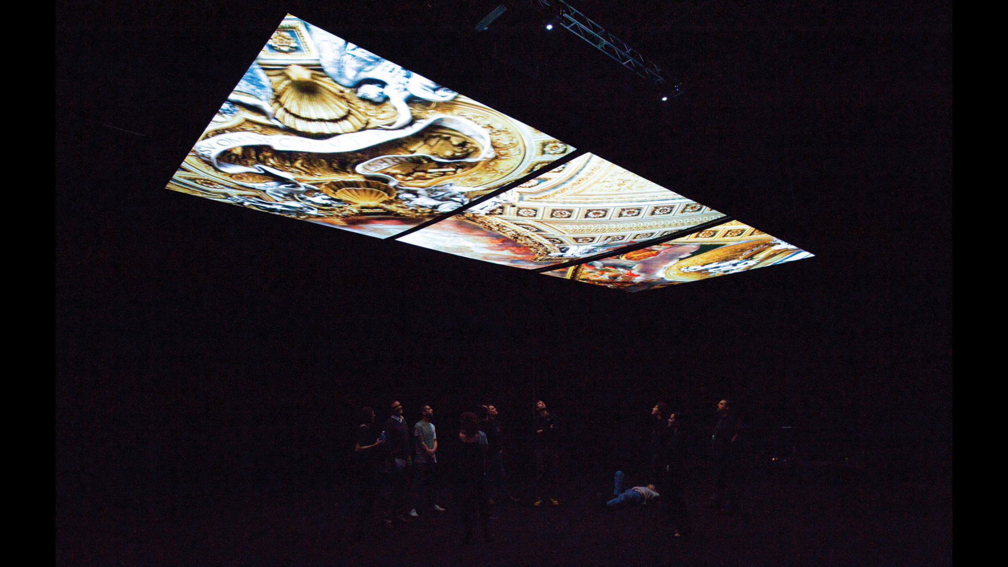 Three screens showing images of an ornate palace ceiling suspended form the ceiling of a dark room as a small crowd looks up.