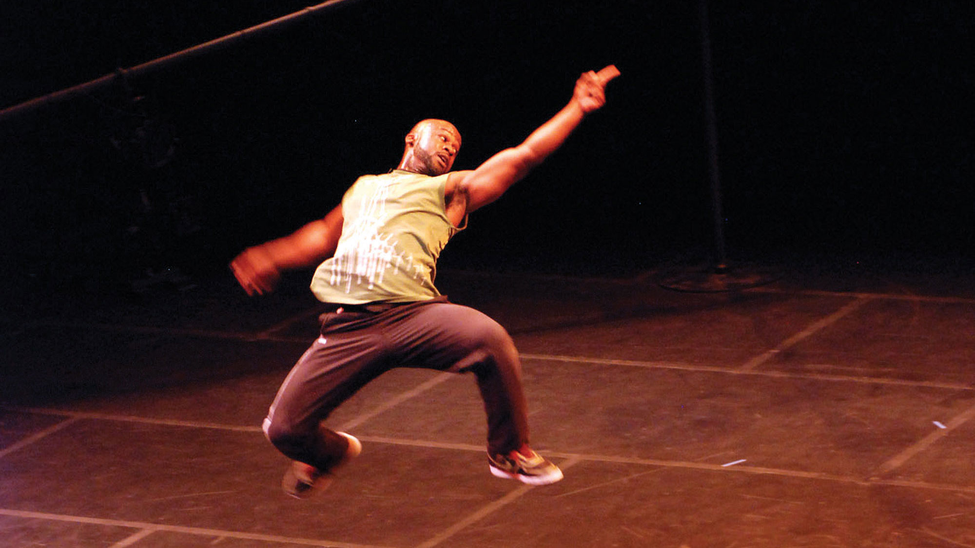 A bald Black man wearing a green tank top and gray pants leaping through the air with back arched and finger pointed I the air. 