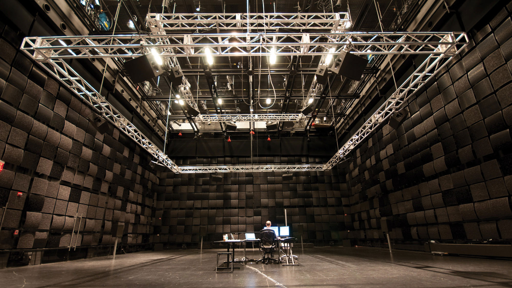 A man seated with back to the viewer at a desk with two putters in a black box studio. A large multure rig is suspended from the ceiling. 