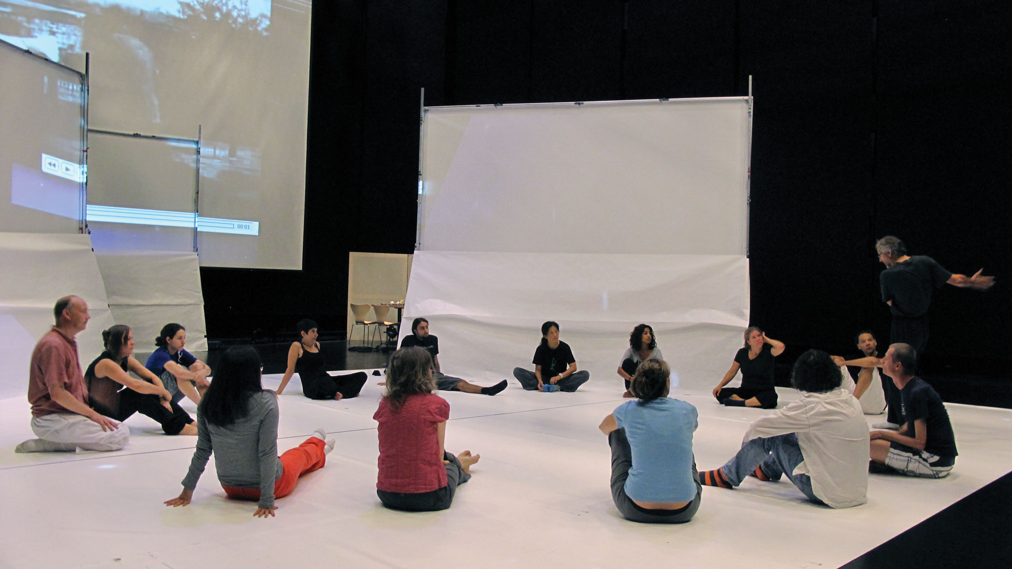 A  group of fourteen dancers dressed causally for rehearsal seating in a circle in a white floor listening intently to a speaker who is standing with arms gesturing outward.