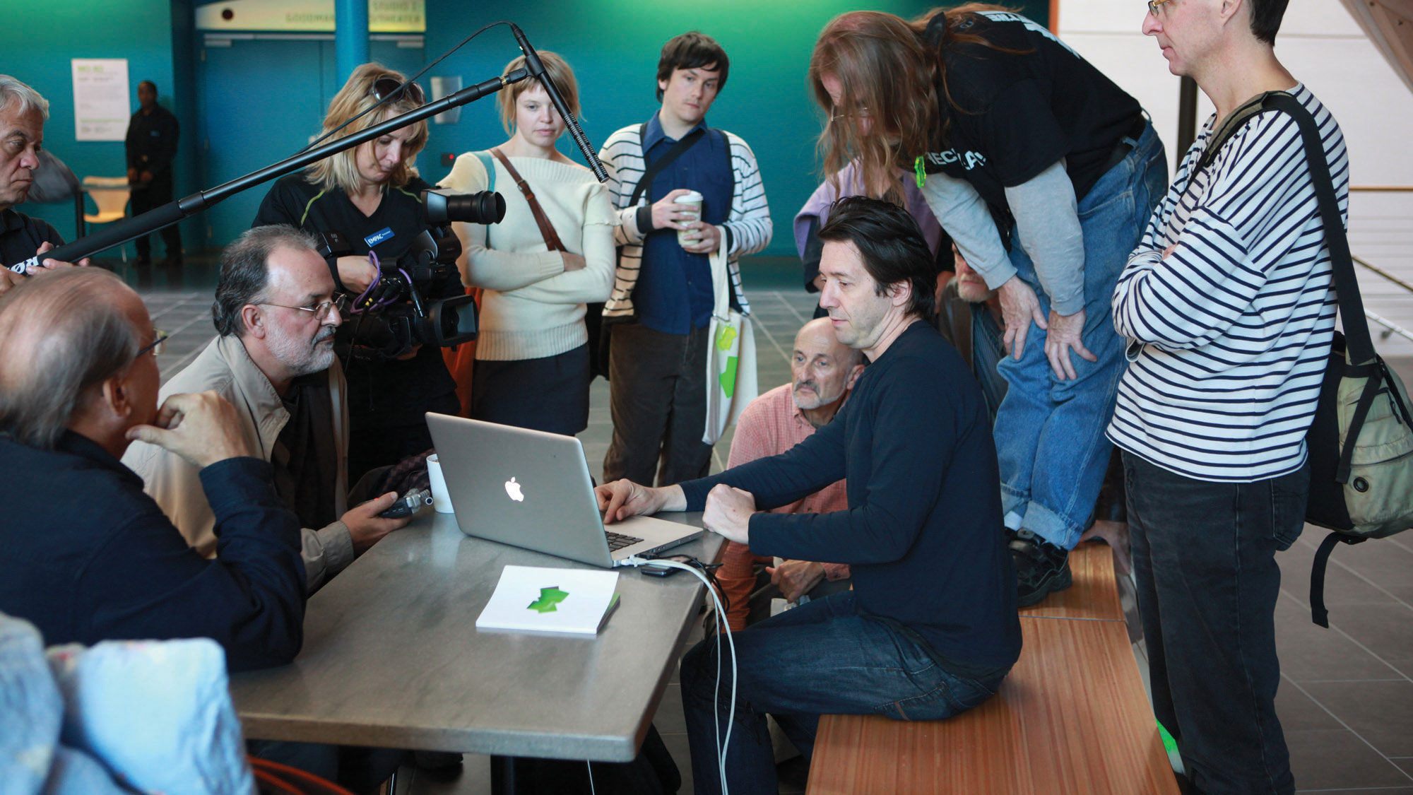 A group of about ten people gathered around a MacBook on a gray table. A white man wearing a black hoodie operates the computer as a small crew of a camera, recorder, and boom mic record. 