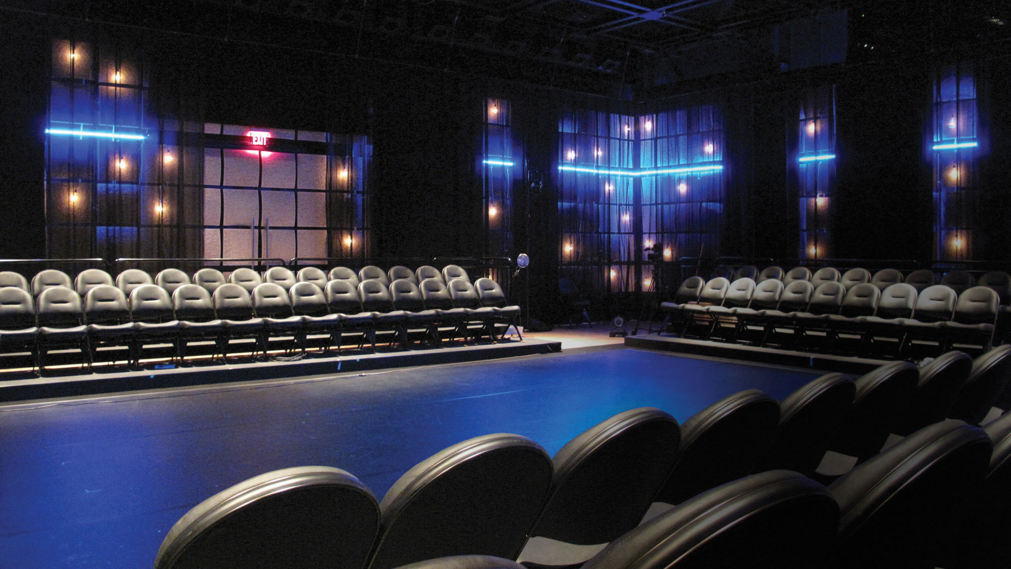 A black box theater lined with black folding chair seating, lit with blue lights in preparation for a show. 