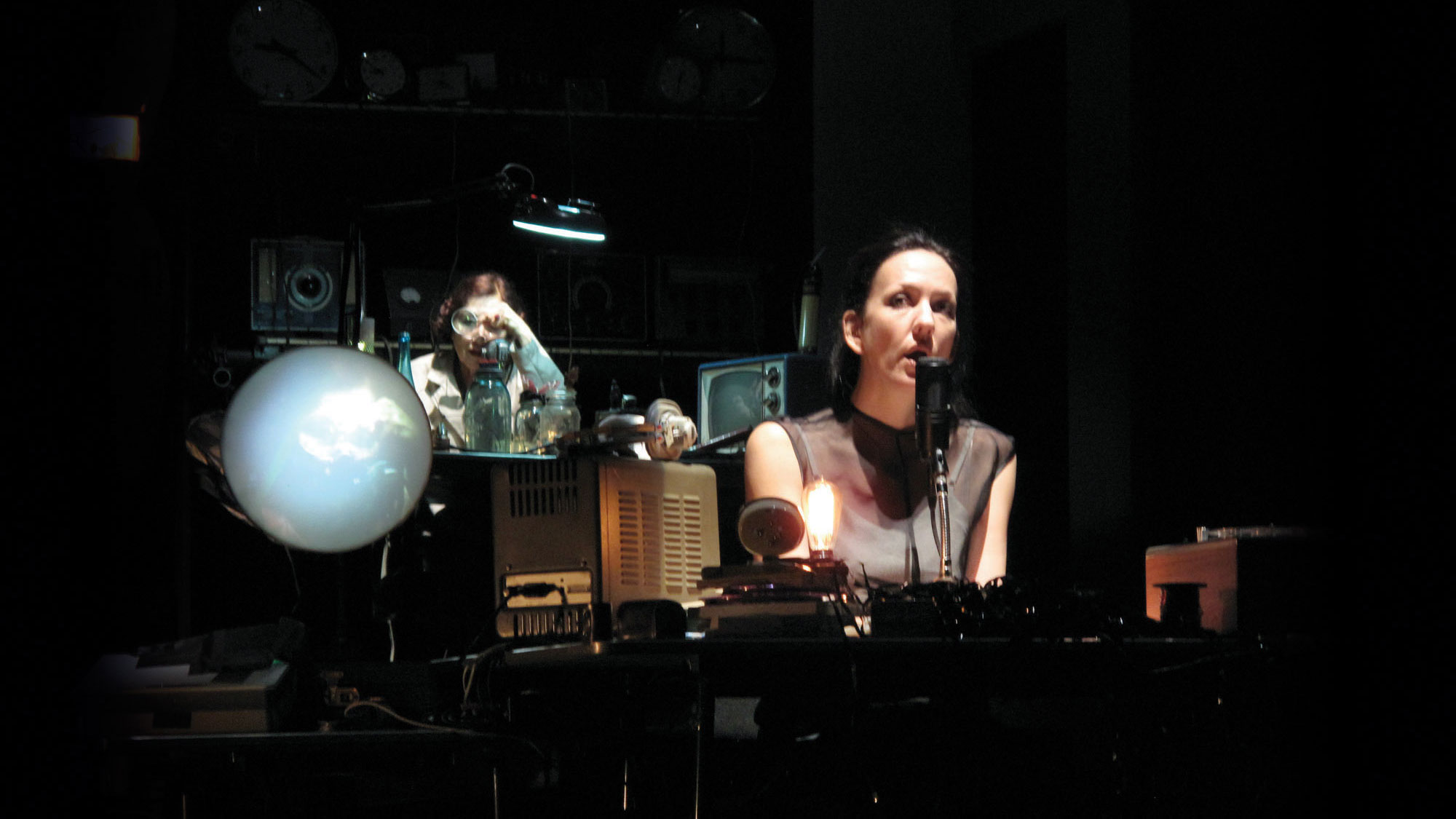 A white woman wearing a brown sleeveless top speaking into a microphone on a dark stage sounded by various vintage sound equipment silhouetted. Another woman with red hair styled in a 50's style sits behind her looking through a magnifying glass at glass jars set in front of her. 