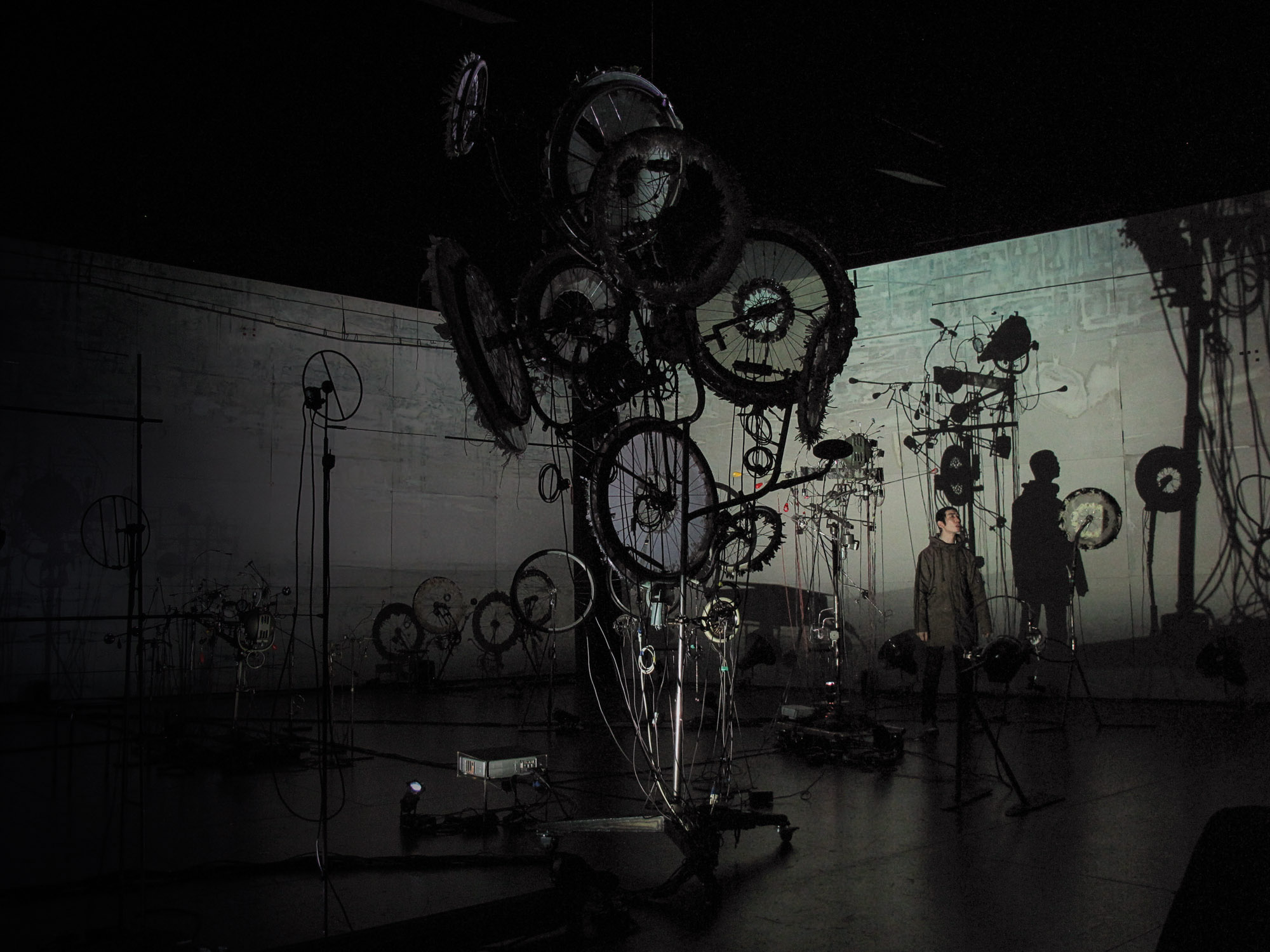 A man casting a shadow standing amongst an abstract sculpture of various wheels in a moody gray room. 