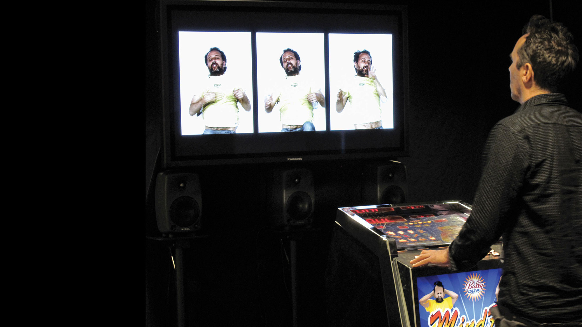 A man playing a pinball machine in front of three projected images of another man in various states of motion. 