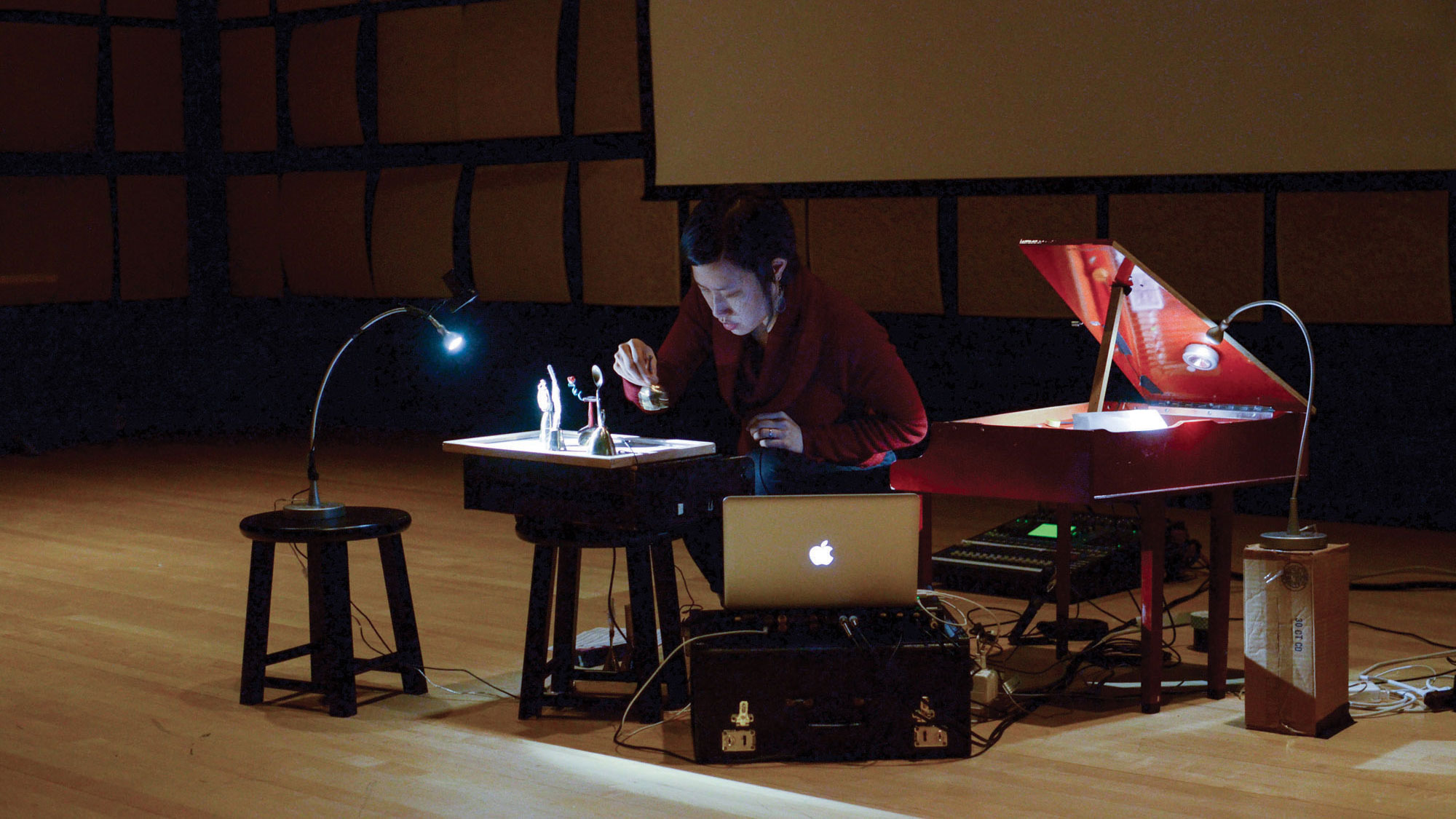 A woman hunched at a small desk setting up figurines under a light. She is surrounded by a small red piano and sound recording equipment. 