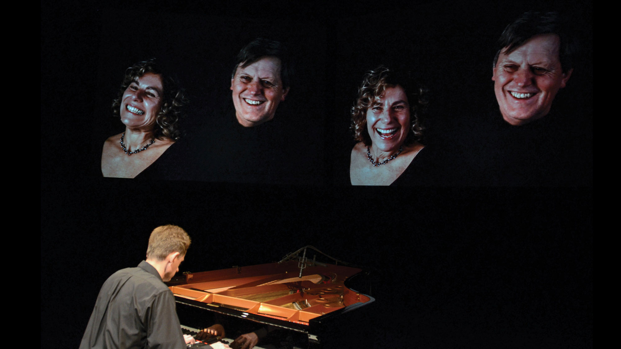 A man seated at a piano with back to the viewer as four projections of smiling people floor above him. 