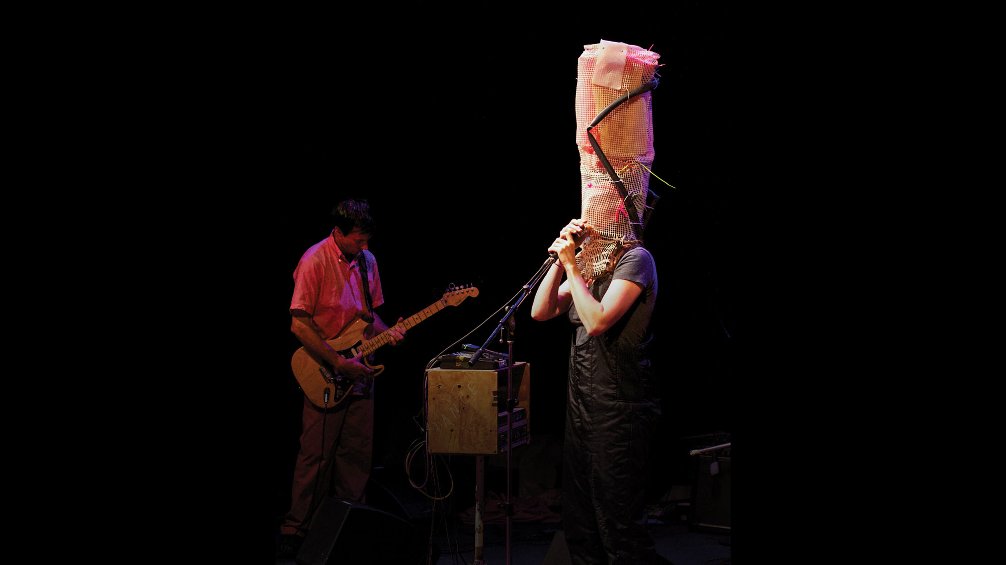 A person singing into a microphone while wearing an abstract tall headdress of wicker canning, zip ties, and a bike tire. 