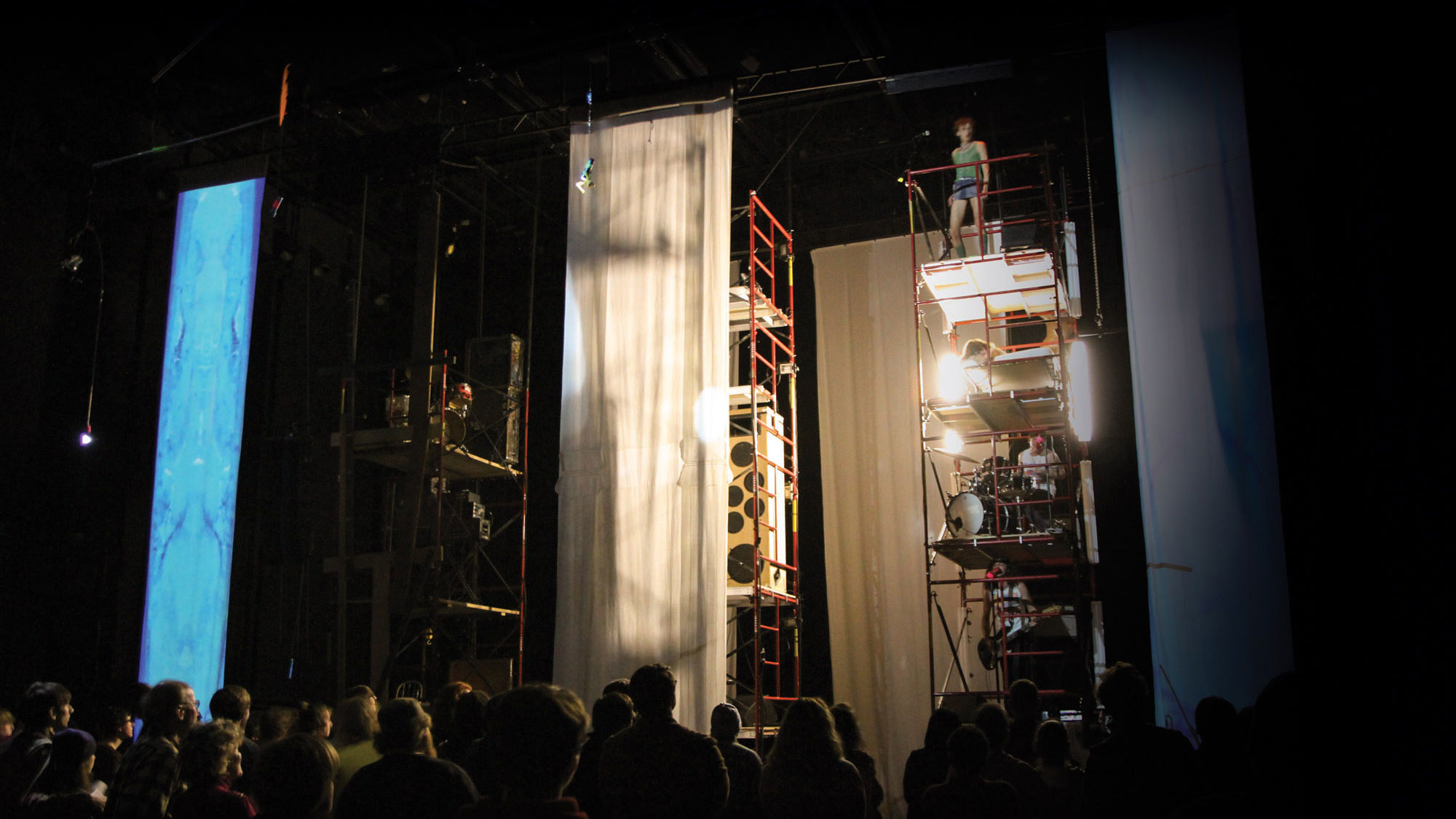 Musicians on scaffolding with each member of the band on different levels playing a concert to a crowd on a staged draped in sheer white fabric. 