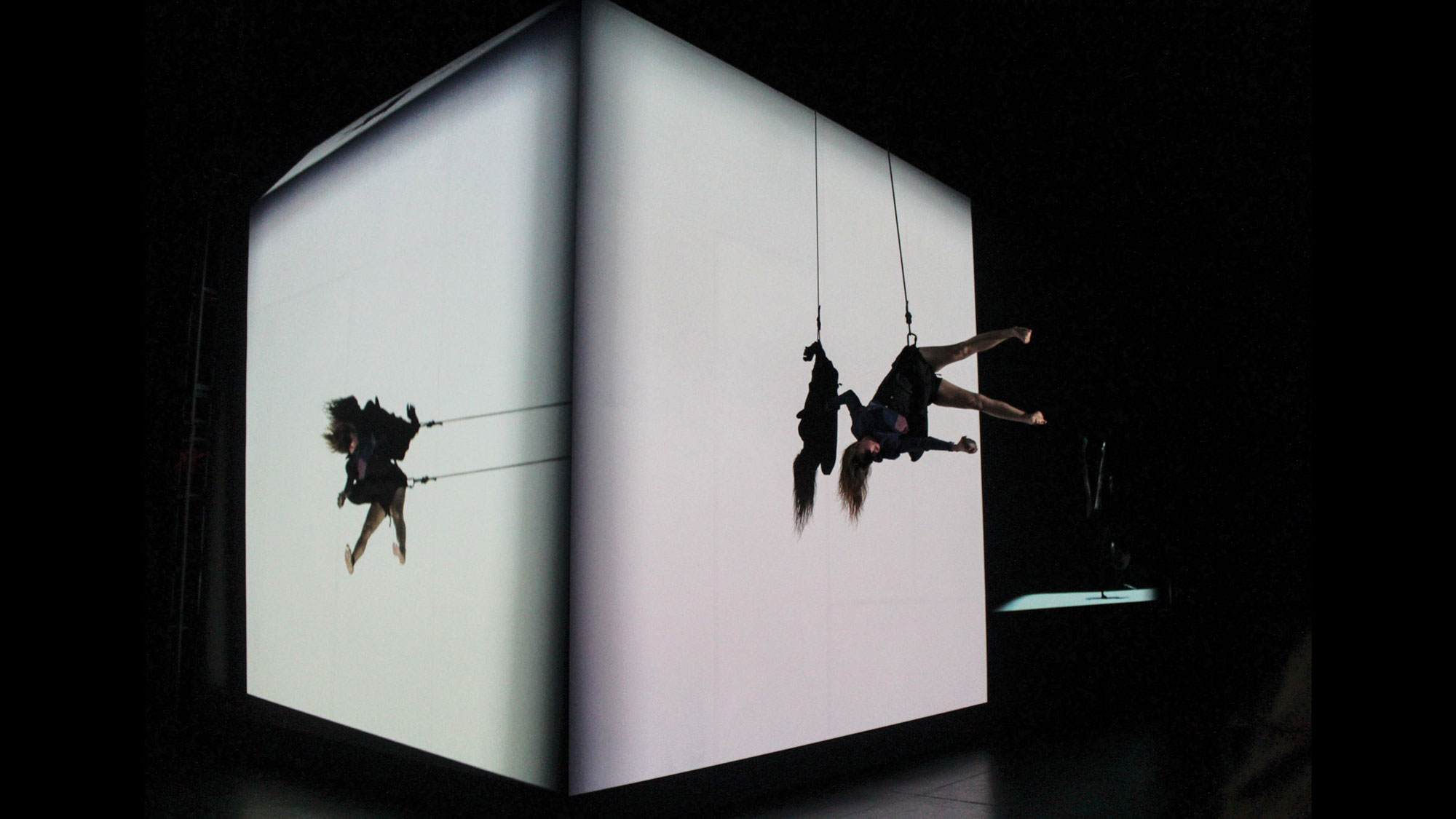 A woman suspended by a cable harness seemingly defying gravity while dancing on a white cube. 