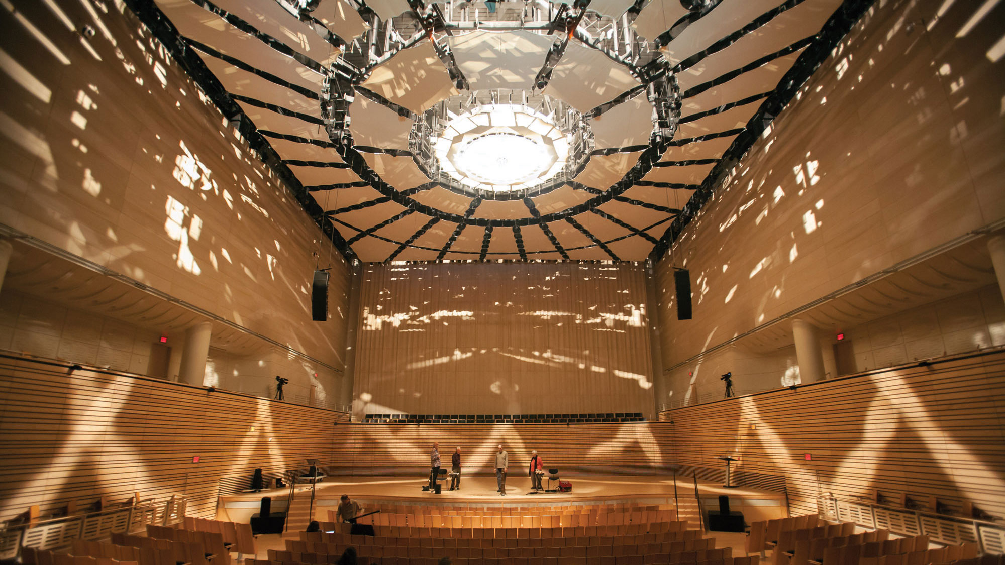 The EMPAC Concert Hall lit with spotty filtered light, mimicking the way light dapples through trees. A group of people stand on the stage among a few chairs. 