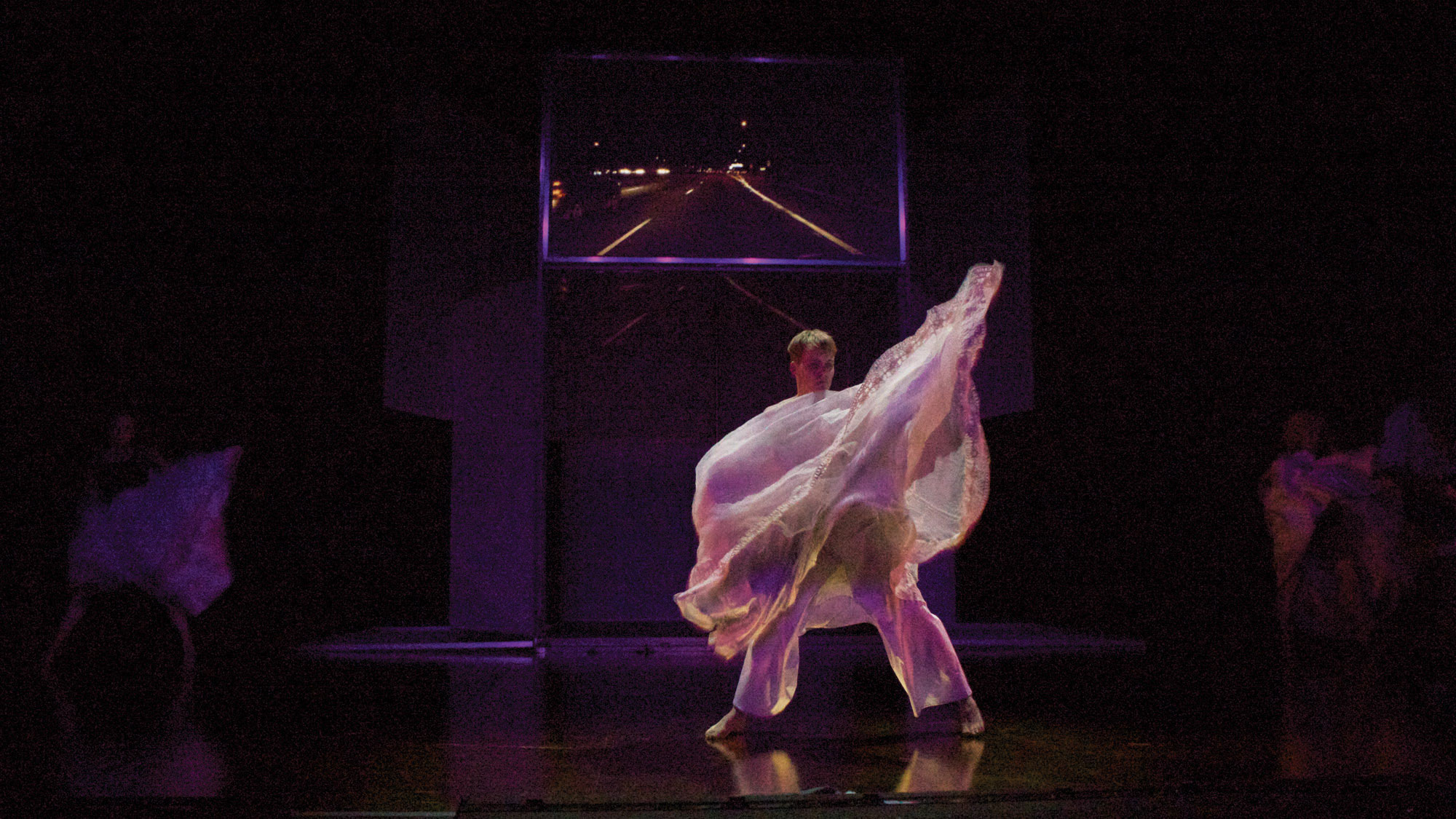 A male dancer wearing a sheer white flowing costume dances with an arm outstretched on a dark purple lit stage infant of a small screen projecting a road. 