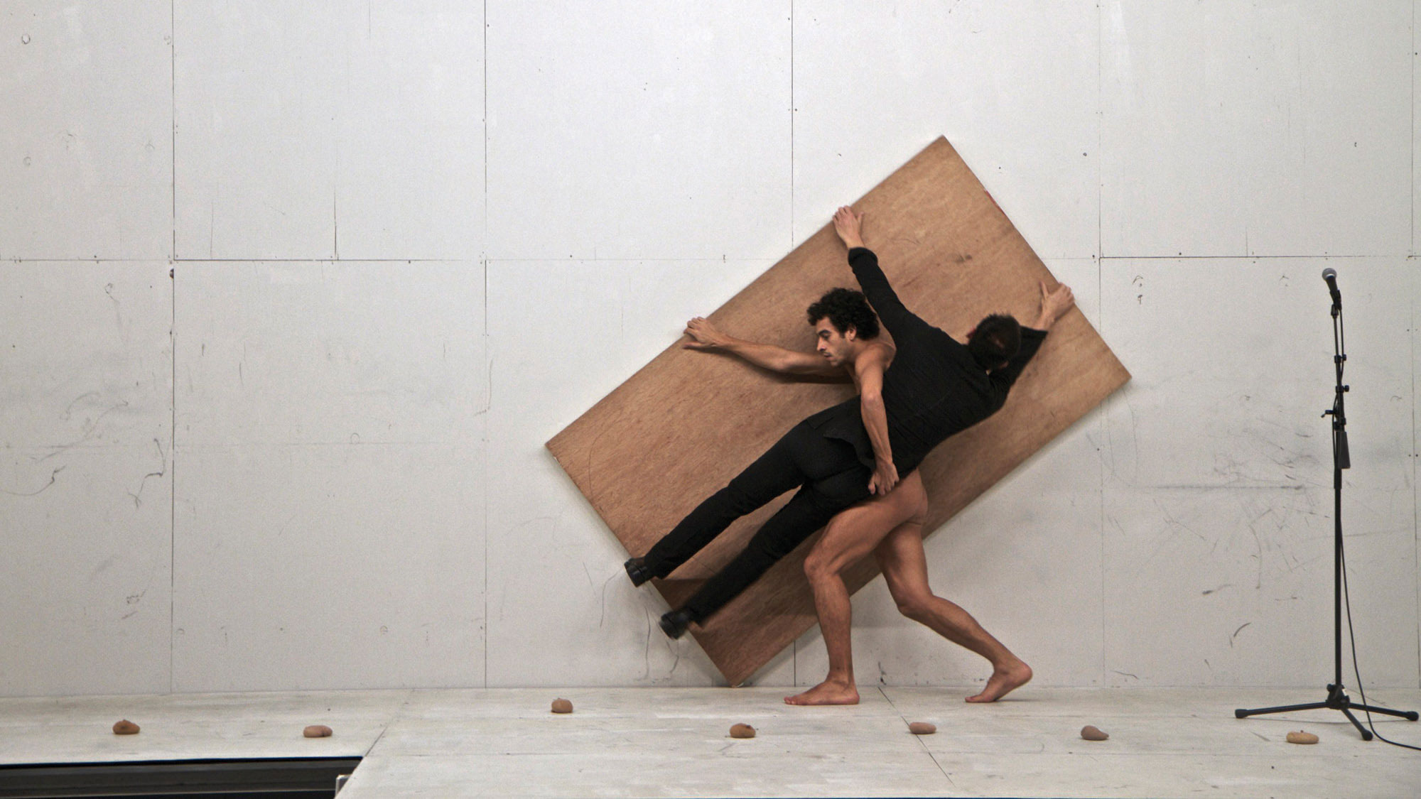 A nude man carrying another man wearing a black unitard while both hold a large piece of plywood on a white stage. 
