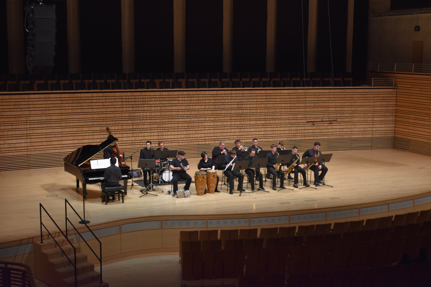 A small orchestra dressed in black on the concert hall stage playing their instruments facing out at the audience. 