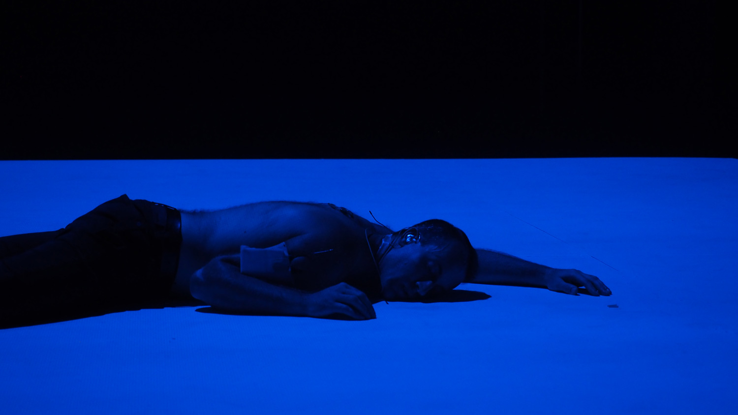 A male performer laying on stage washed in blue light. 