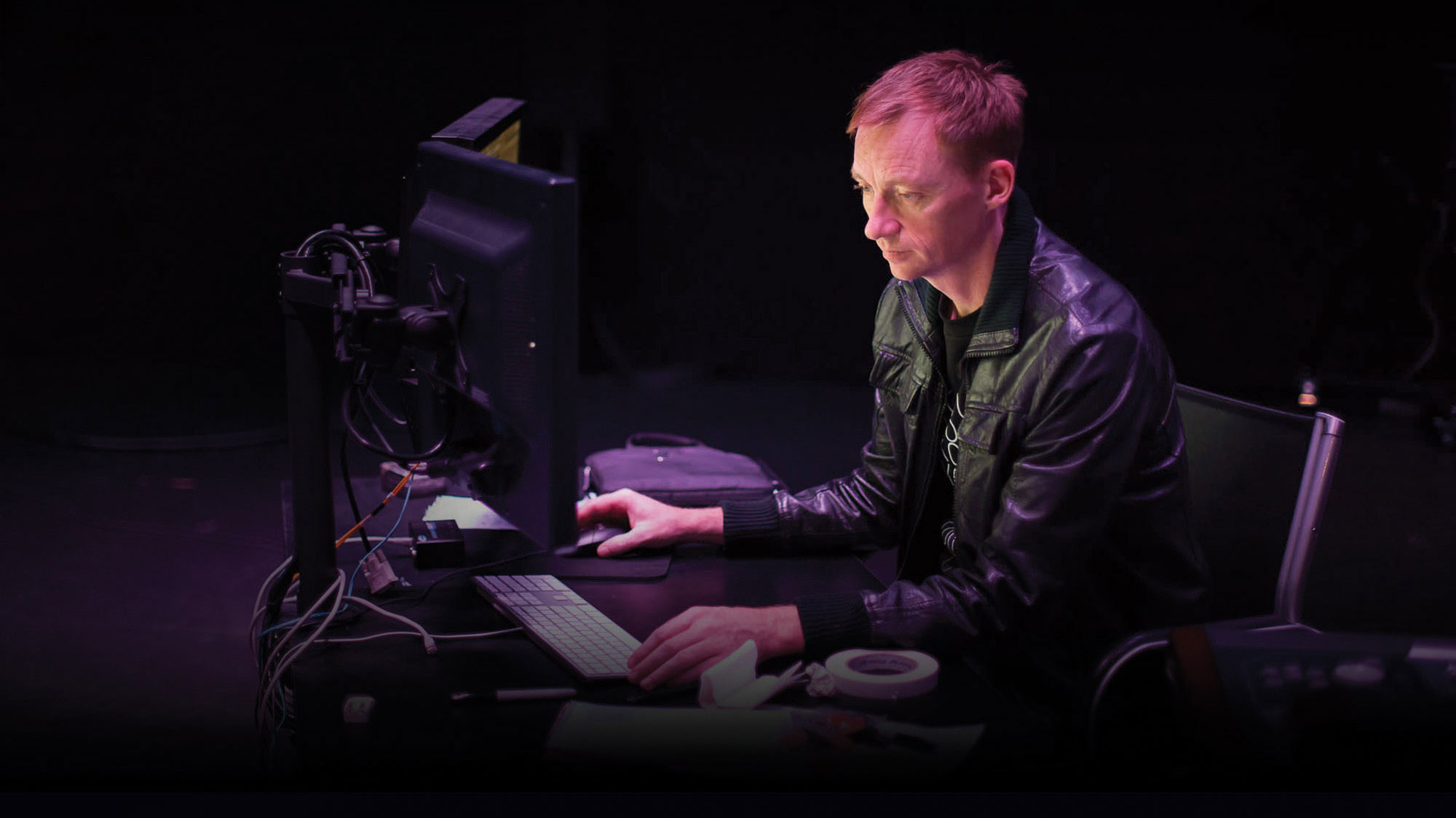 A white man wearing a leather coat seated in front of a dual computer console. 