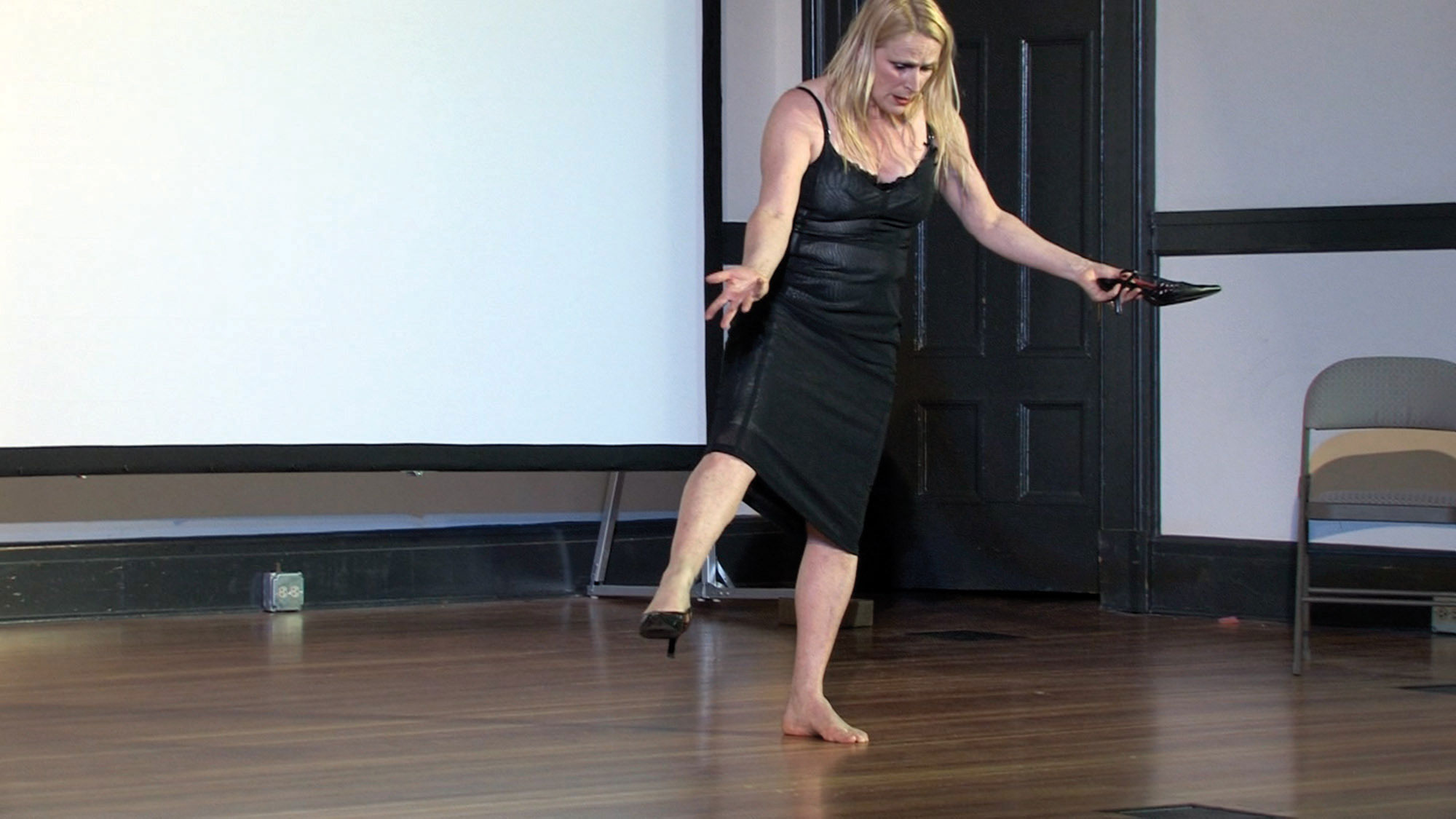 A femme dancer wearing a black slip dress and one black heel standing on the shoeless foot with the other leg extended and clutching the missing black heel. She looks down with hands out to the side. 