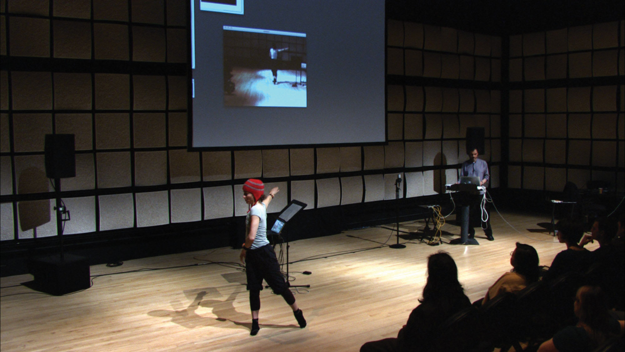 Frédéric Bevilacqua giving a lecture in front of a wall of gray acoustic tiles as a female dancer wearing a red winter hat moves across the stage.