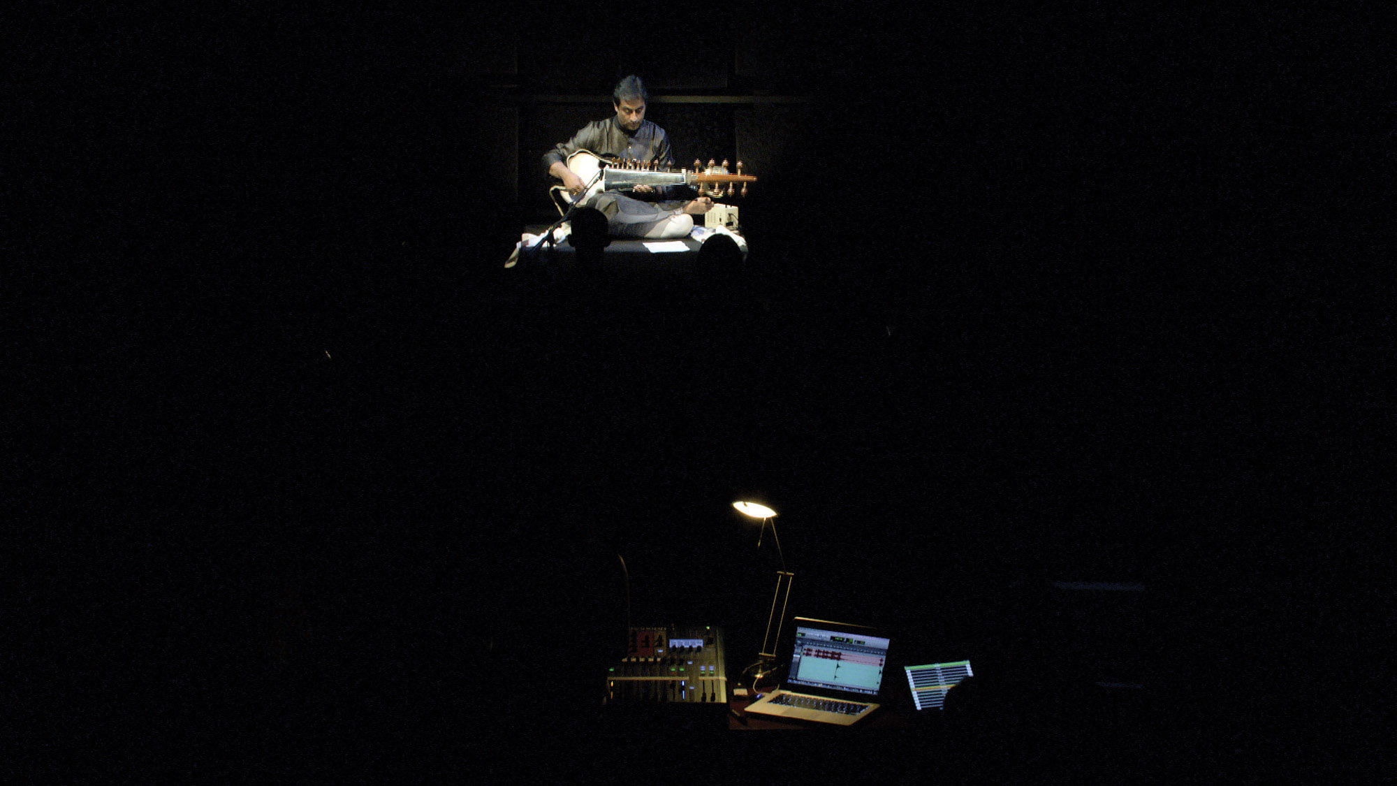 A man seated playing the sitar in a dark room, lit only by the sound panel in front of the stage. 