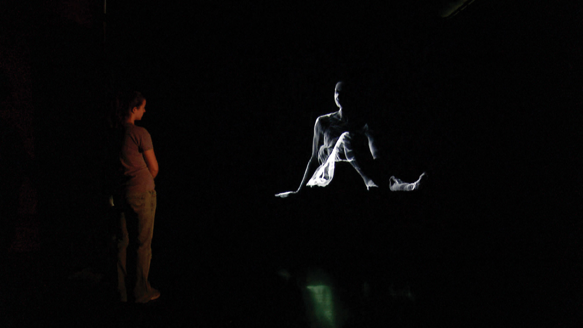 A woman studying the projected image of a ghostly woman seated with legs out, supported by her arms behind her. 
