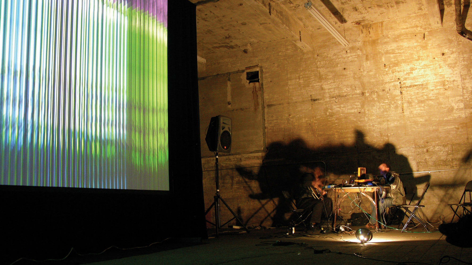 Two blurred people sitting at a folding table with various equipment and wires in an industrial looking space with an abstract projection of green, purple, and blue lines. The pair is lit from below causing long shadows. 