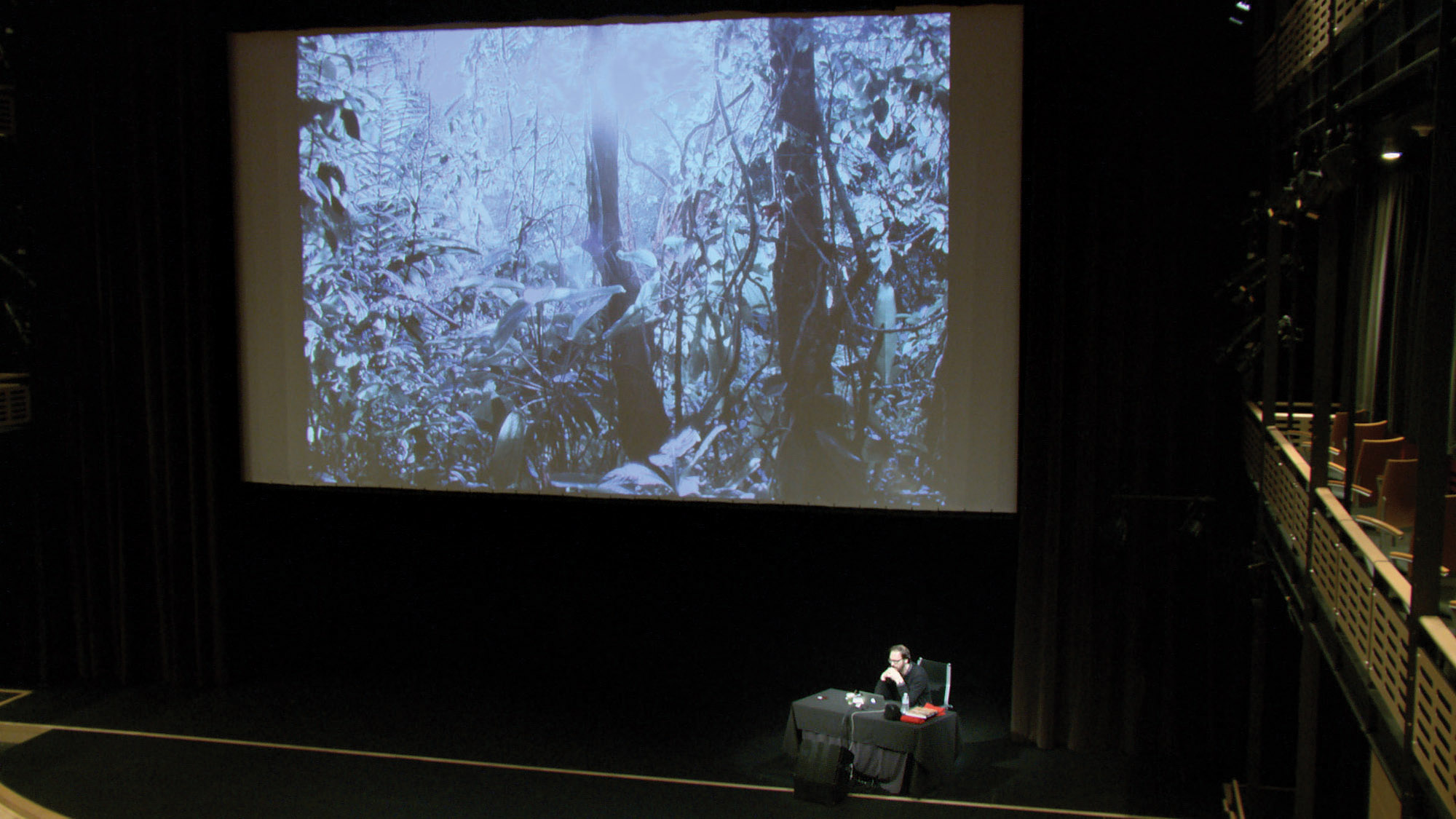 A man sits behind a table with a black table cloth on stage in front of a large screen projecting an image of jungle foliage. 