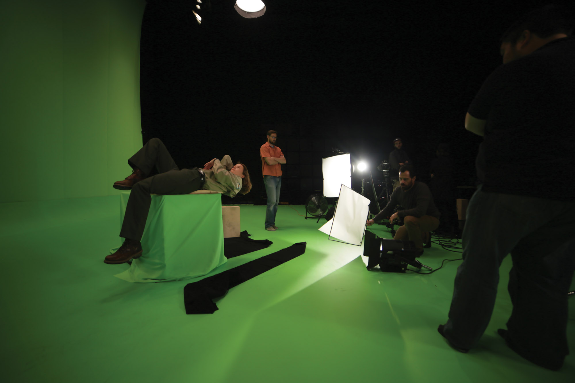 Five people doing various production tasks work around a woman laying on a green cube in green room. 