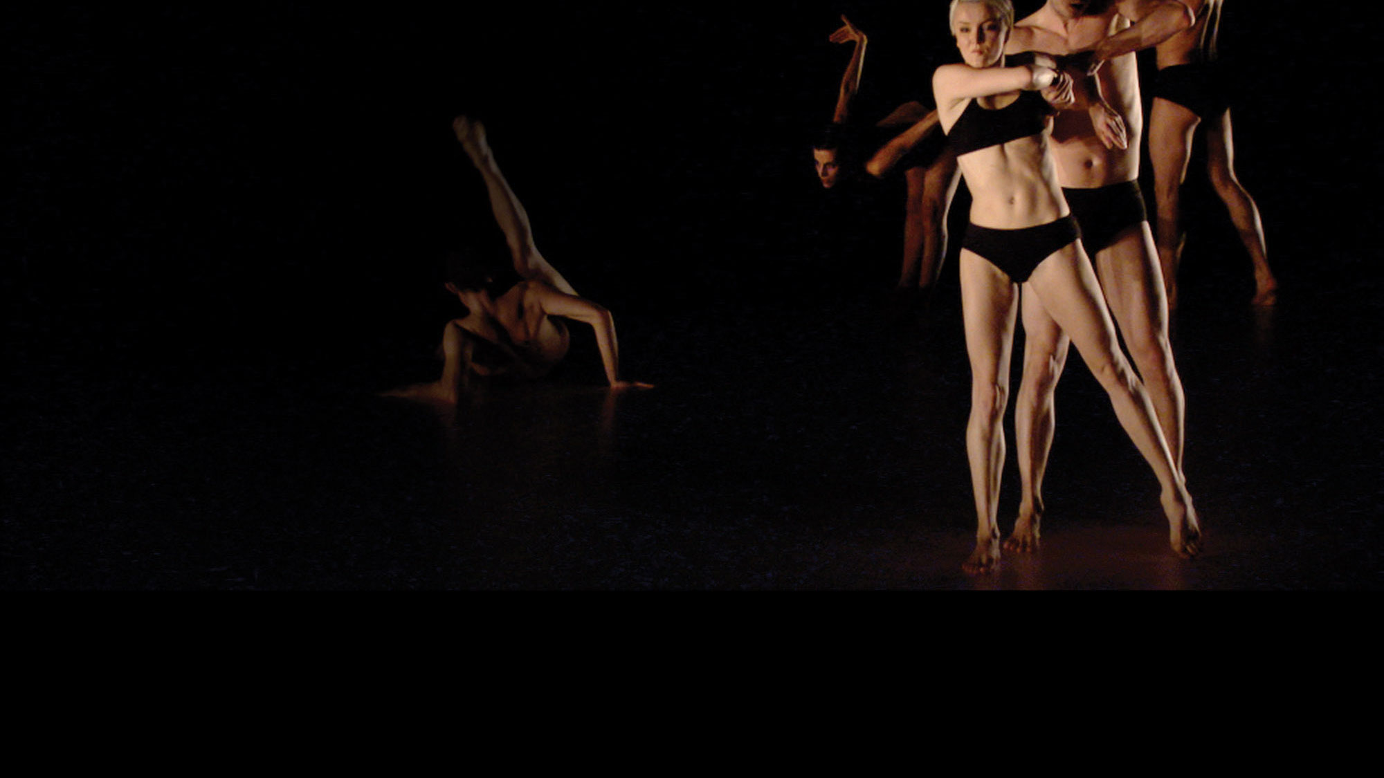 Five dancers wearing black underwear preforming on a black stage. A male dancer can be seen on the ground, back to the viewer with leg up as the rest are in a diagonal line. 