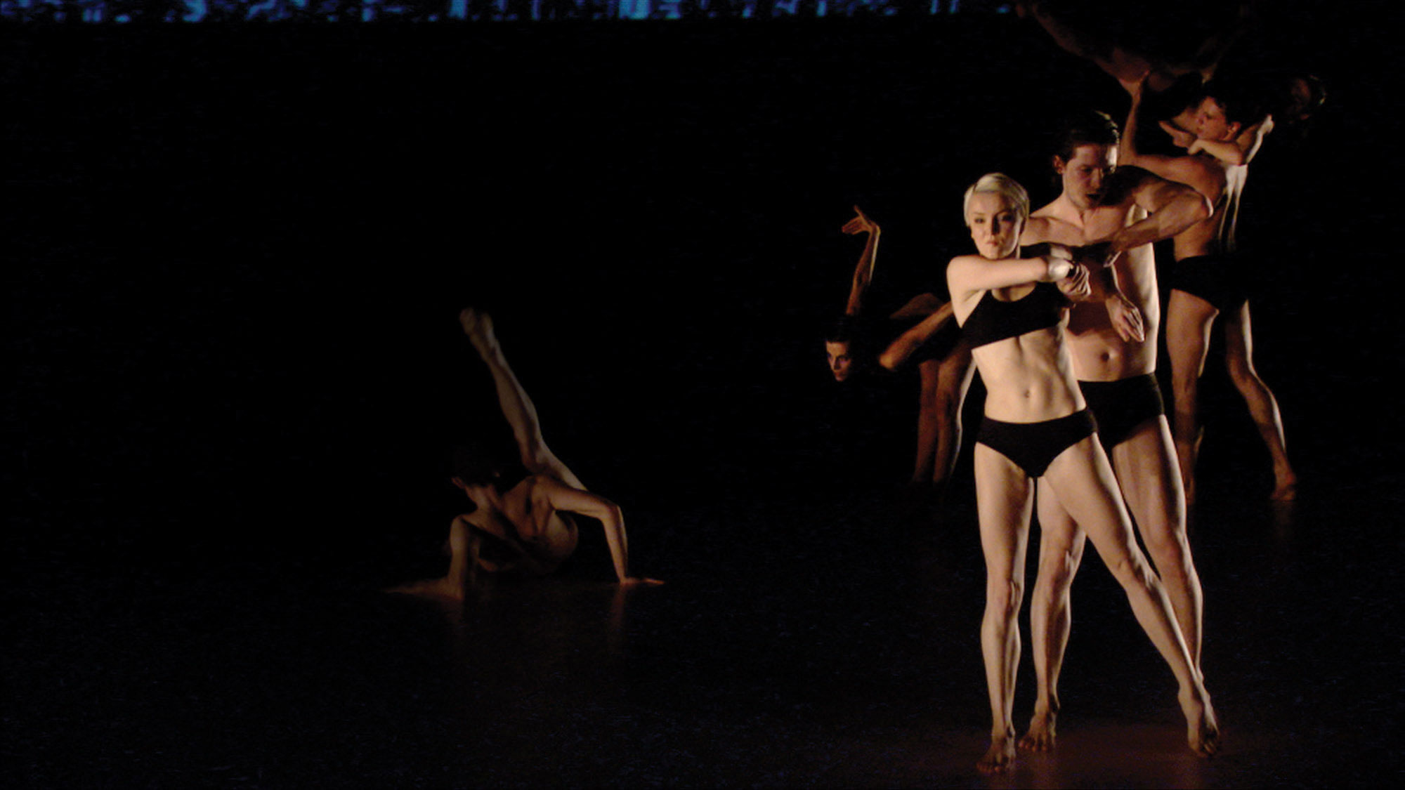 Six white dancers on dark stage dressed only in black underwear. Each person is in there own unique pose, including an individual on the floor with their leg extended and a woman standing in the front with arms twisted. 