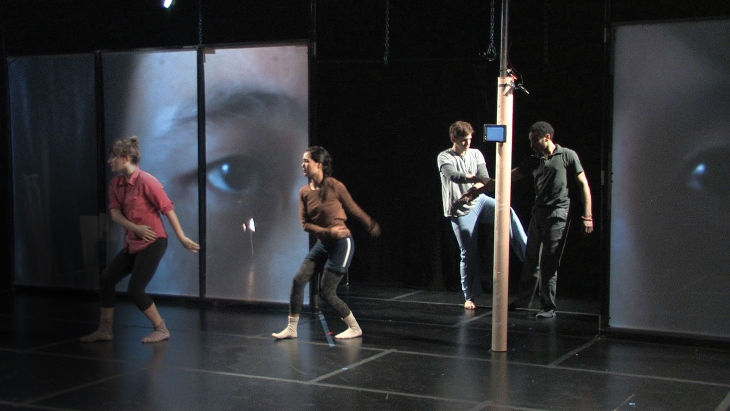 Four dancers wearing street clothes dancing in front of a large projection of a human eye on a black stage. 