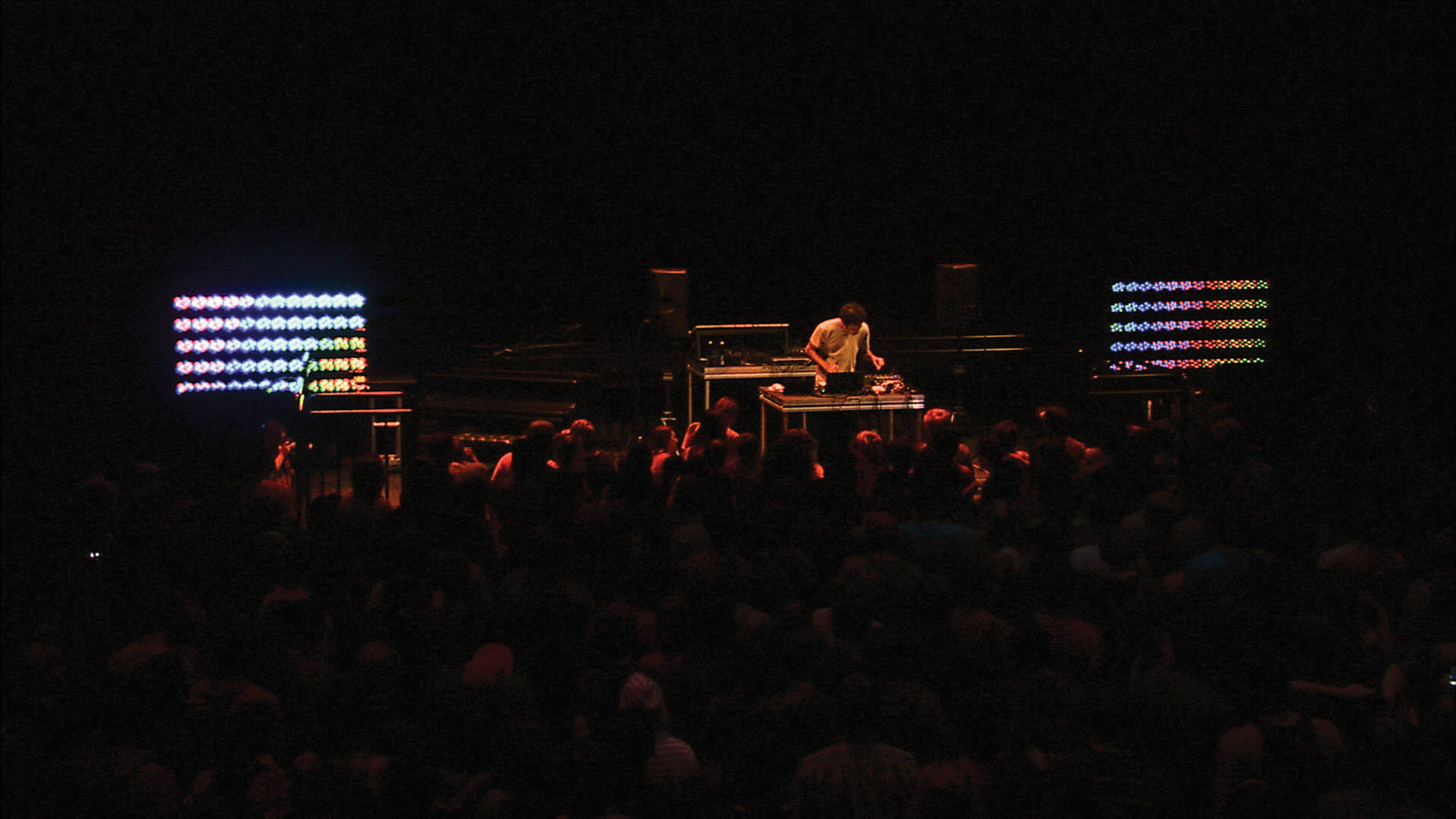 A man DJing to a packed crowd in a dark room lit only by dim red light and the two screens on either side of the stage showing lines of rainbow shapes. 