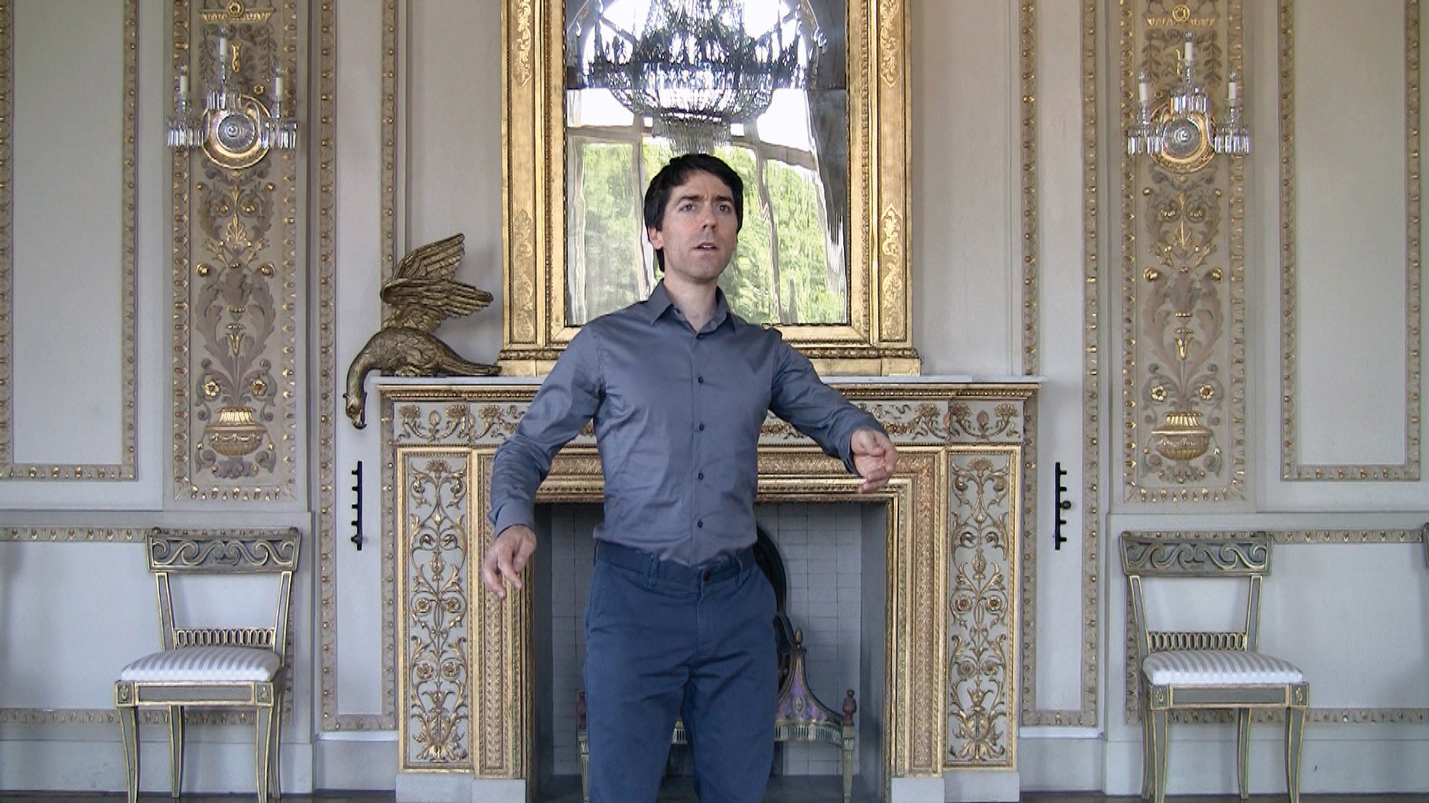 A white man with dark hair wearing a blue button up and pants standing with arms gently gesturing outward in a baroque styled room. 