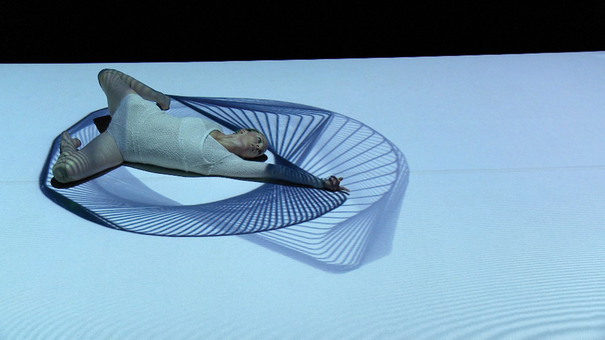 A female dancer wearing a white leotard laying on her side with arm outstretched and legs bent on a spirograph projected onto the floor. 